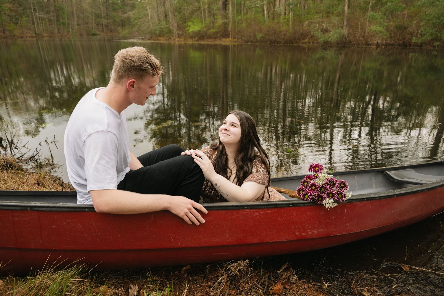 Young couple sitting together in a red canoe on a calm forest lake during engagement session.