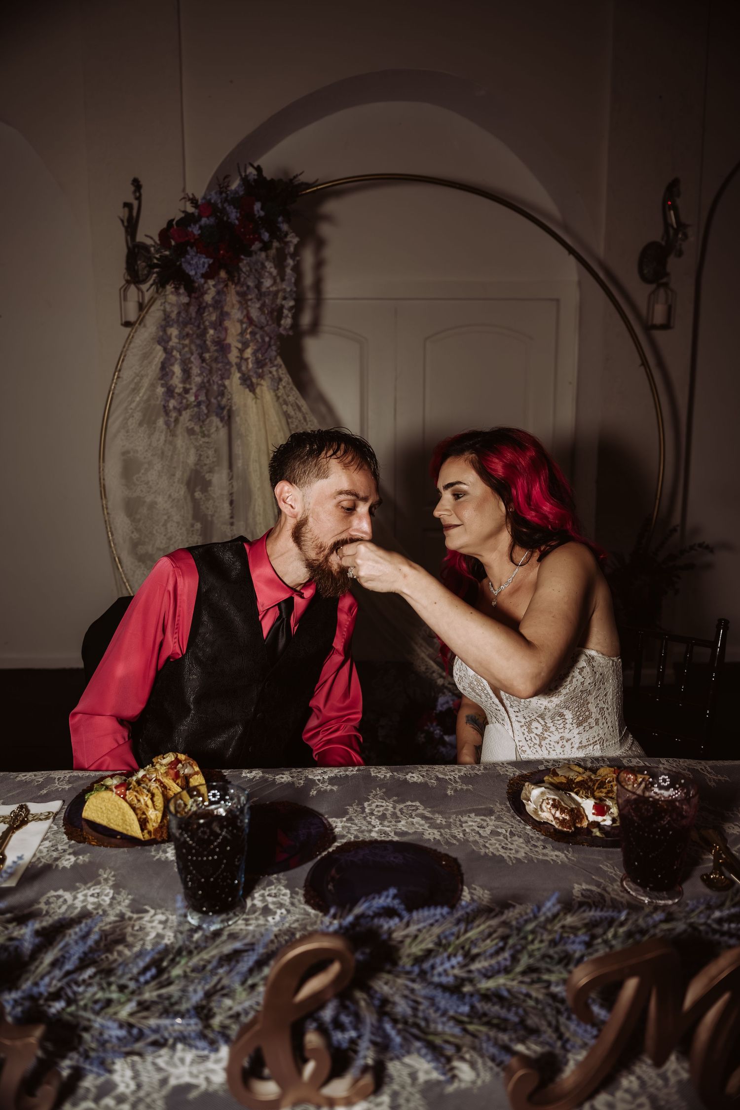 Bride feeding groom at a decorated sweetheart table with floral arch and lavender centerpieces at reception.