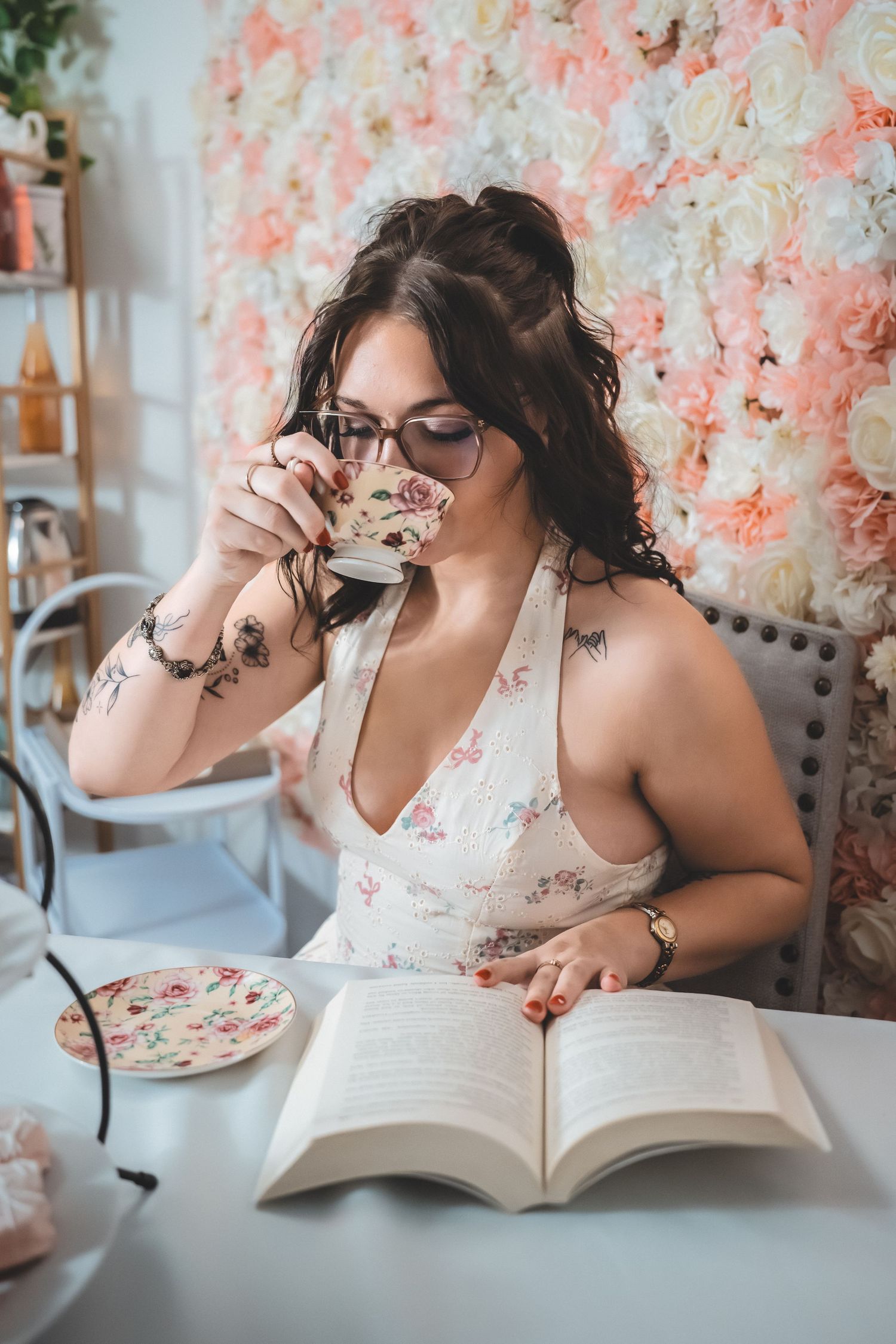Person in white floral dress sits at table with book and coffee against pink floral backdrop in cozy setting.