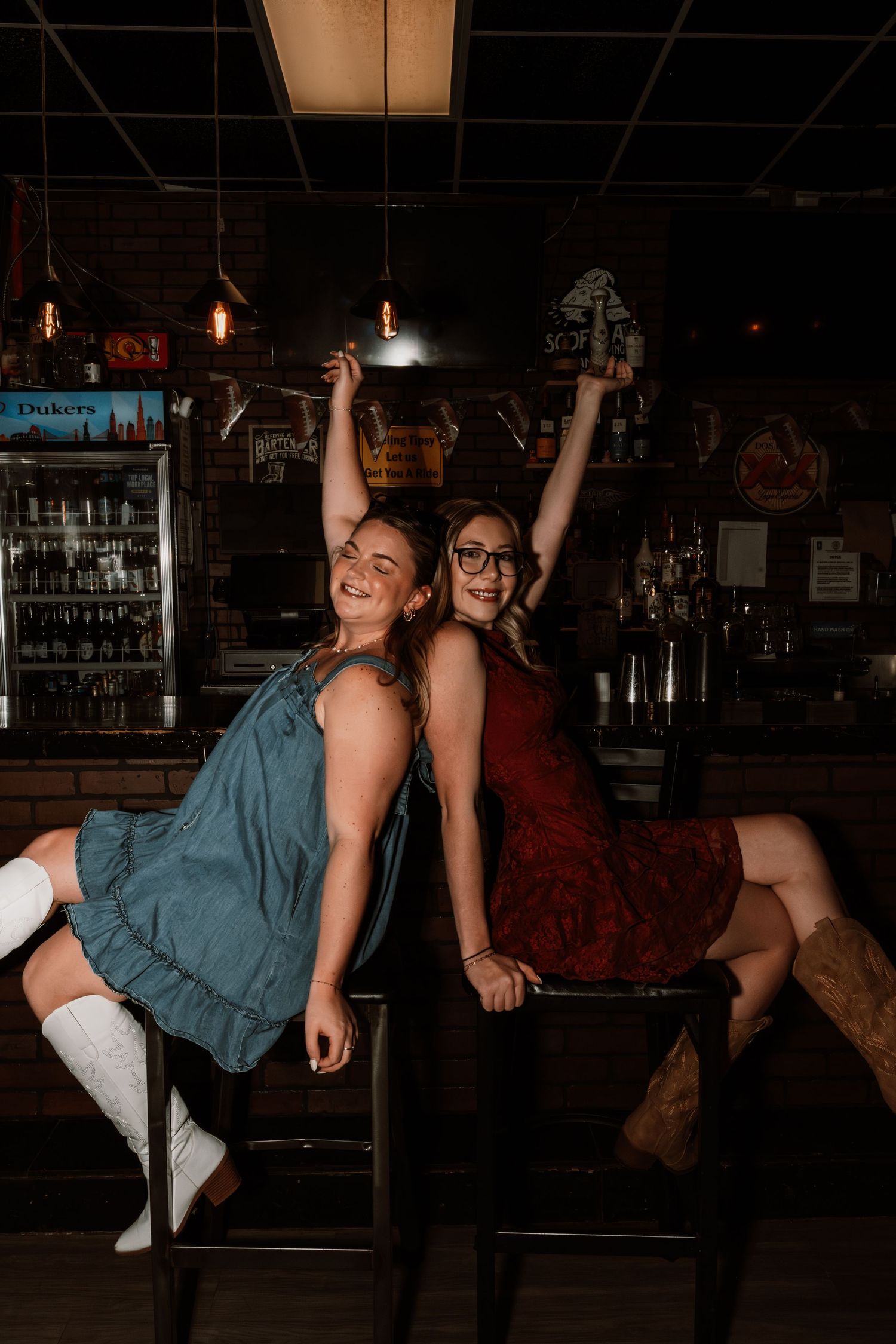 Two smiling women sitting back-to-back on bar stools with arms raised in a bar.