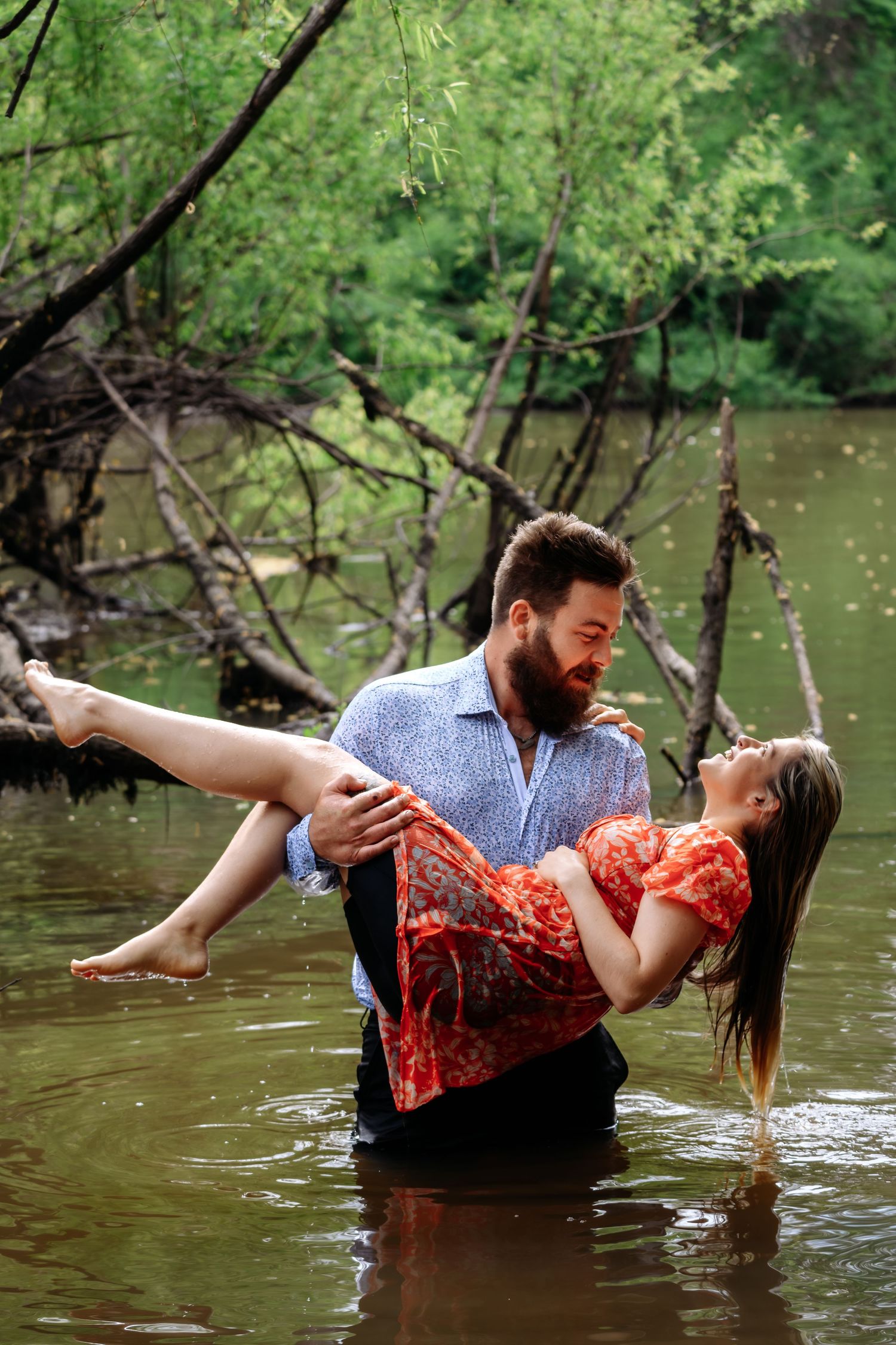 Bearded man in blue shirt carries smiling woman in red floral dress in his arms while standing in a river.