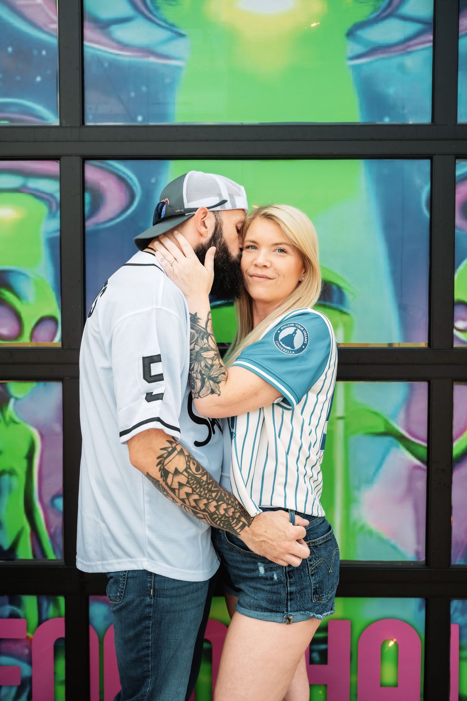 Bearded tattooed man and blonde woman embracing romantically before vibrant alien mural backdrop.