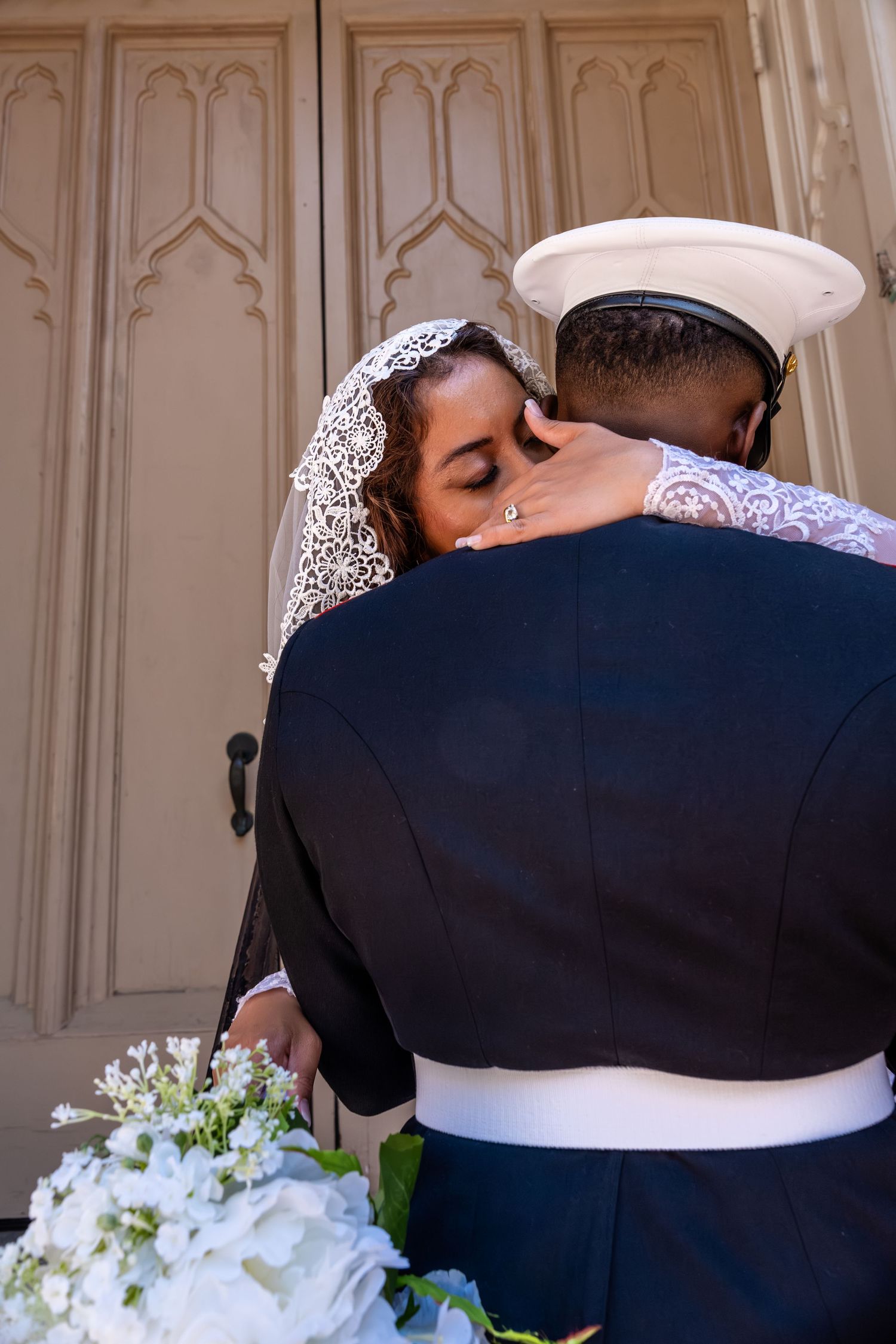 Bride and groom in Marine dress uniform share tender embrace outside church doors on wedding day.