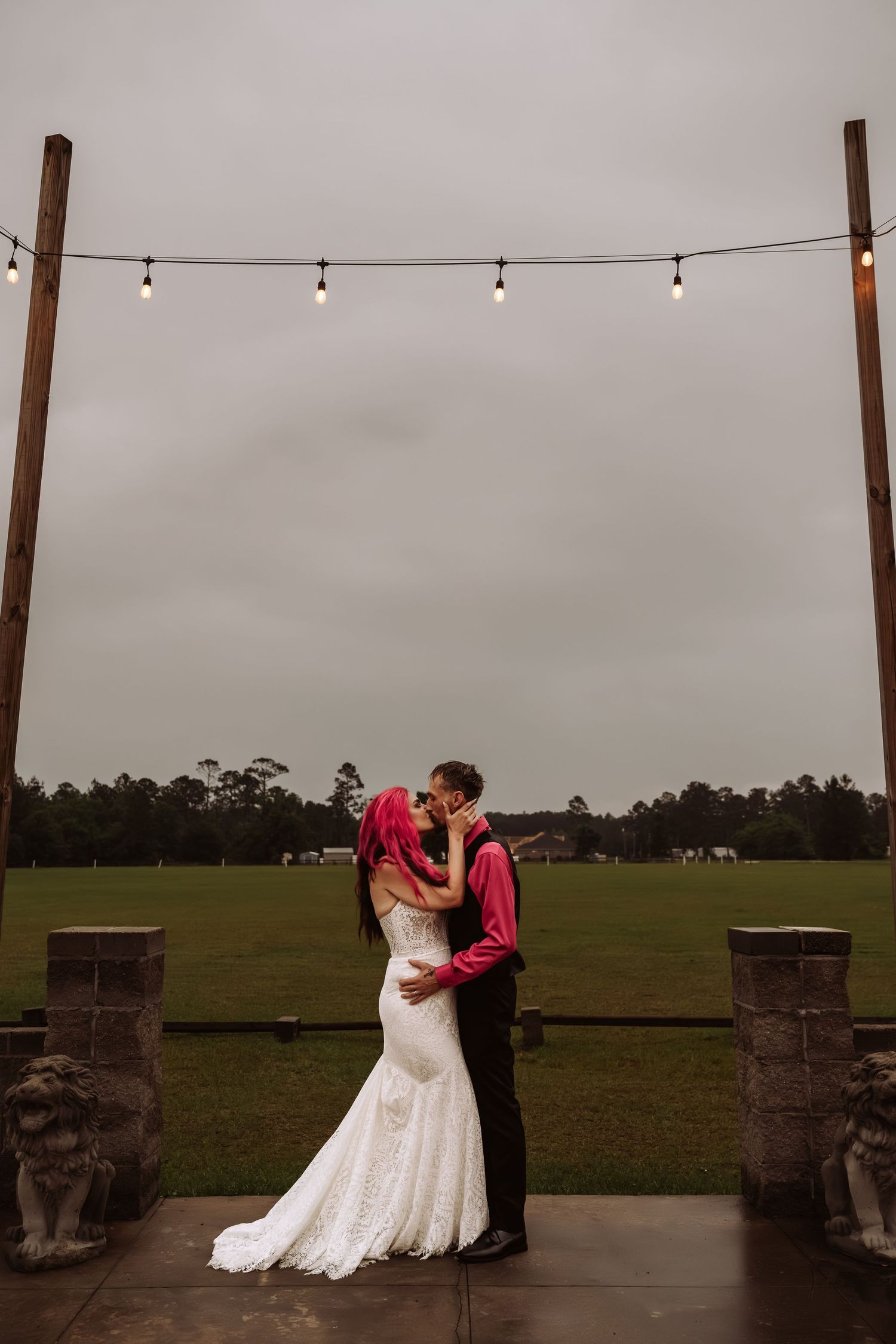 Bride and groom kissing under string lights on an outdoor patio with a green field backdrop.