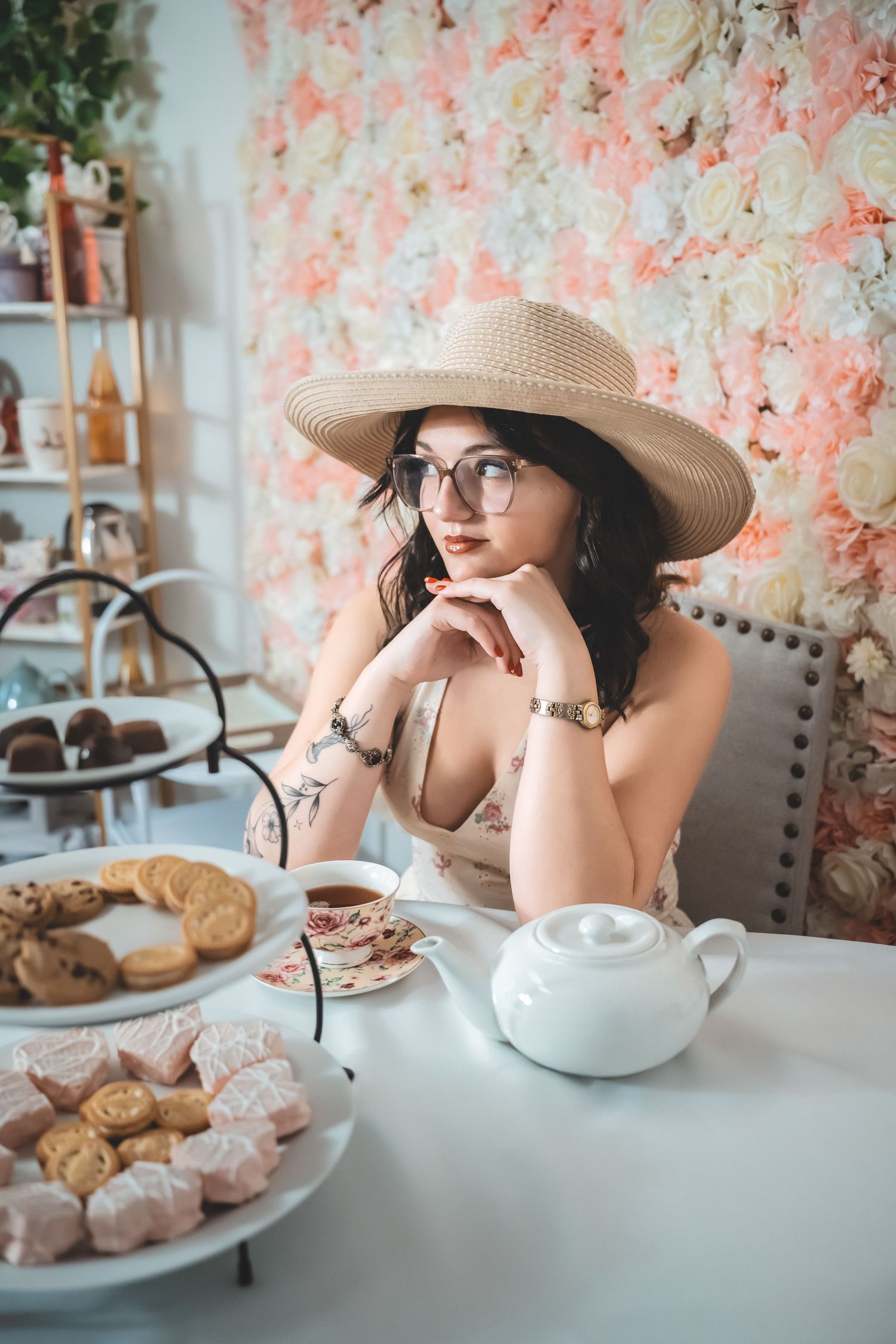 A tea party setting with pink floral backdrop features pastries, teapot, and someone in a sun hat enjoying afternoon tea.