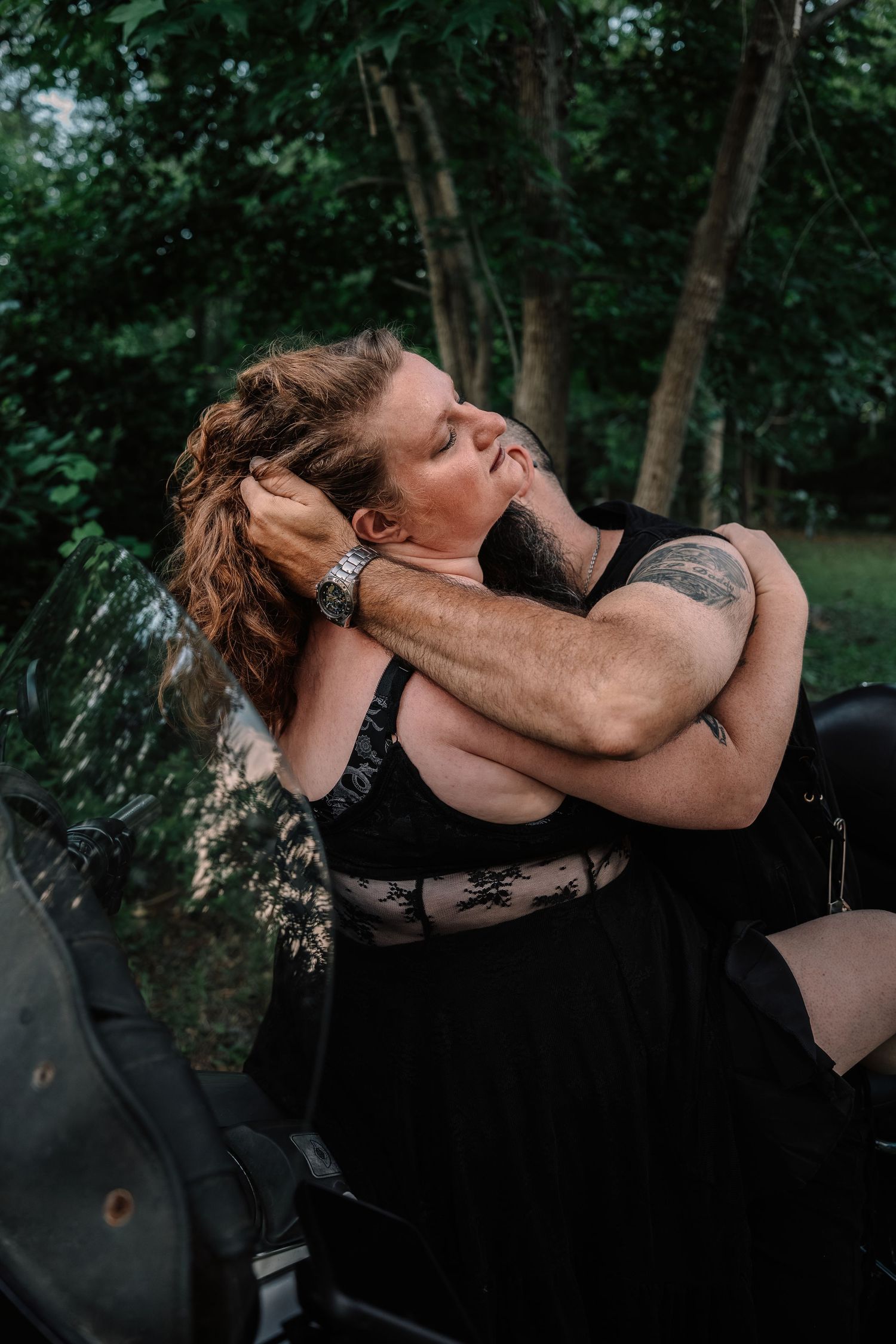 Tattooed couple sharing a kiss beside a motorcycle surrounded by tall green trees.