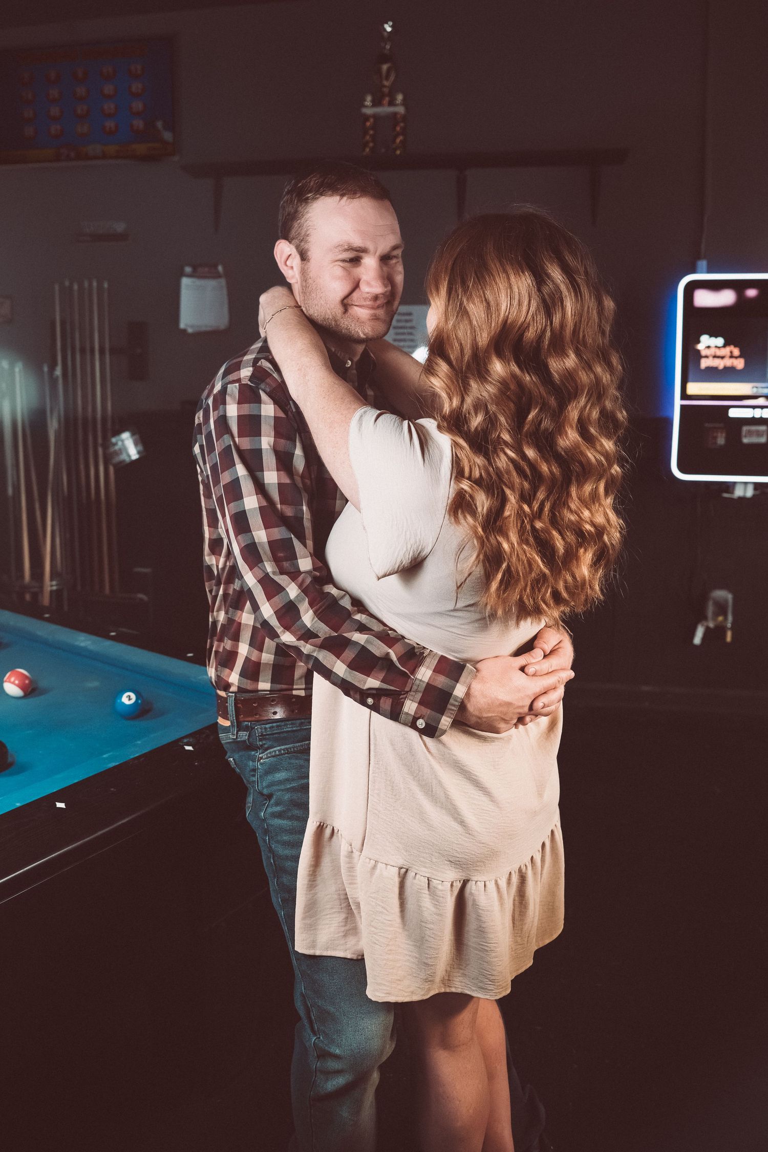 A romantic couple embracing by a pool table in a dimly lit game room with neon signs.