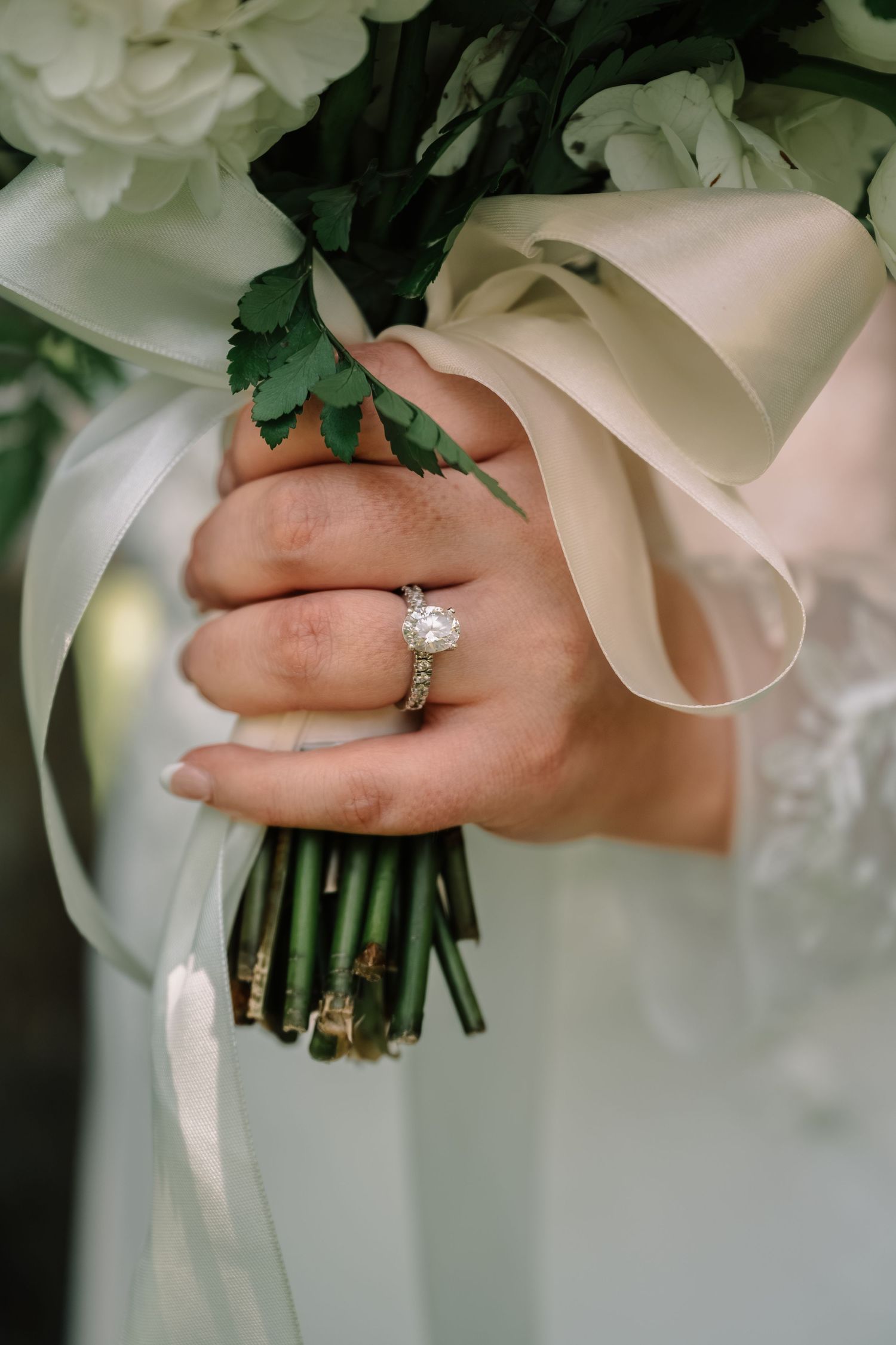 Close-up of a diamond solitaire engagement ring on bride's hand holding a white floral bouquet with ribbon.