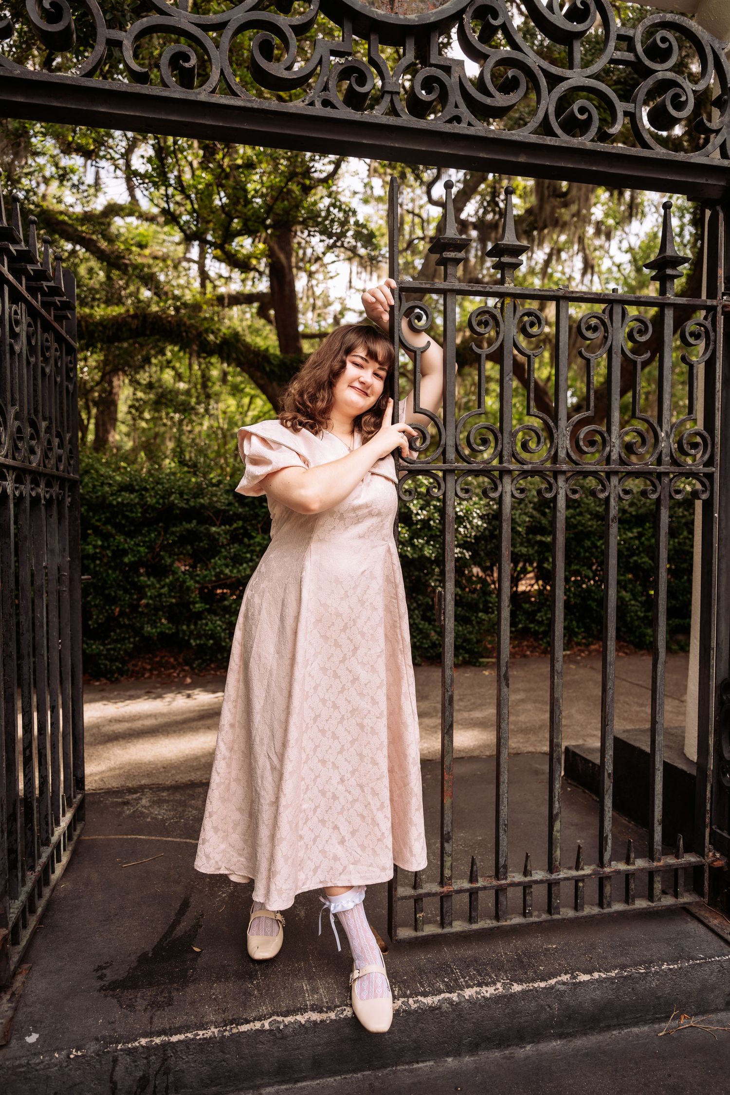 Young woman in blush pink vintage dress posing at ornate iron gate in a garden park.