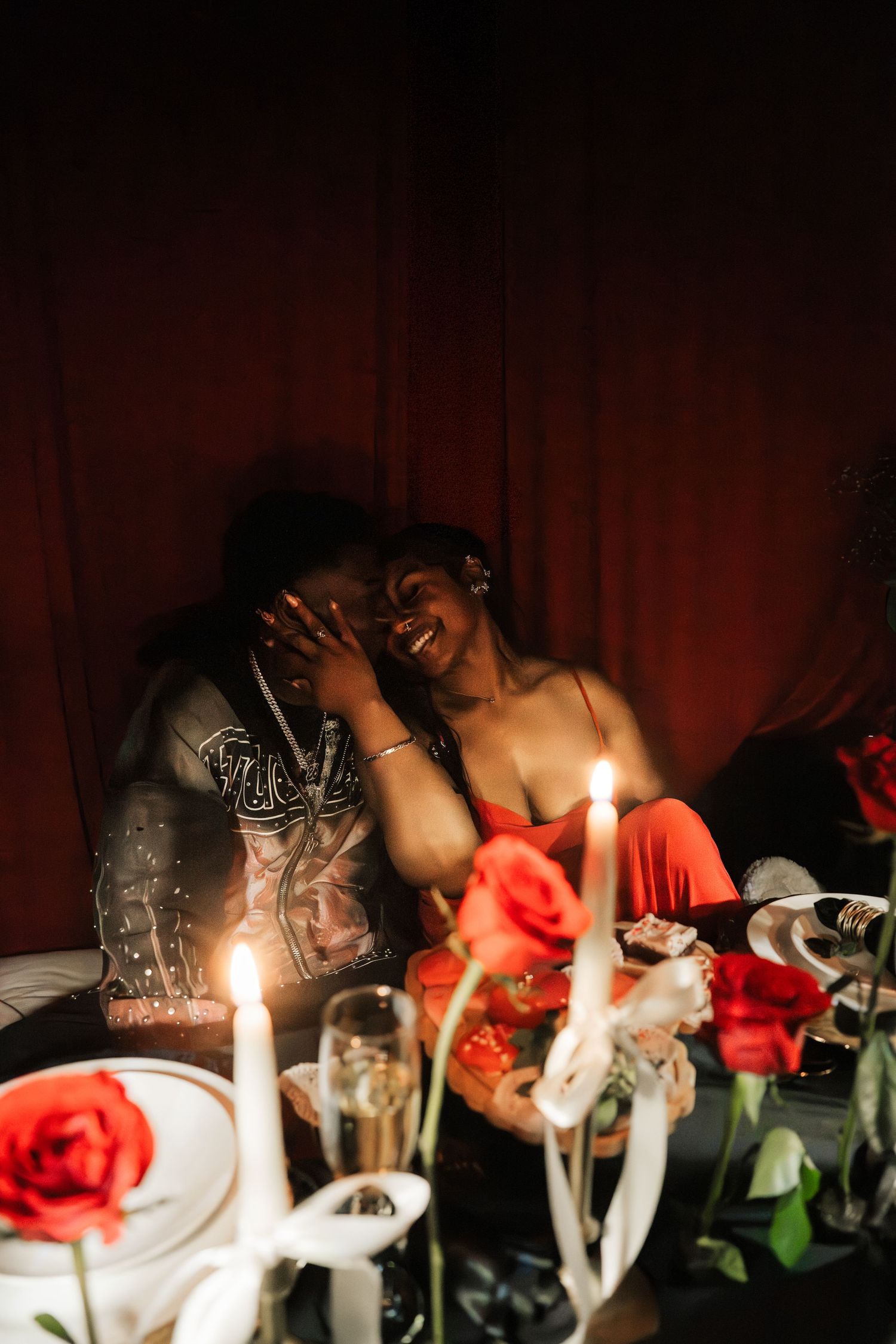 Couple laughs together by candlelight with red roses and champagne in intimate romantic setting.
