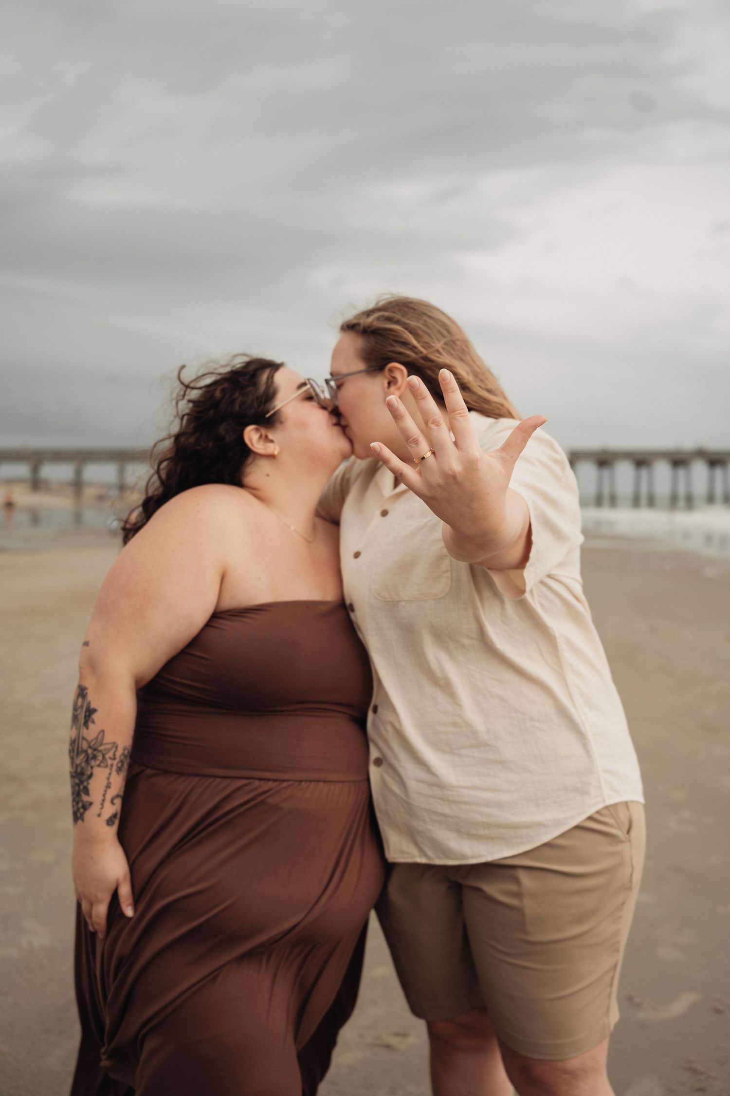 Two women sharing a kiss on the beach while one shows off her engagement ring to the camera.