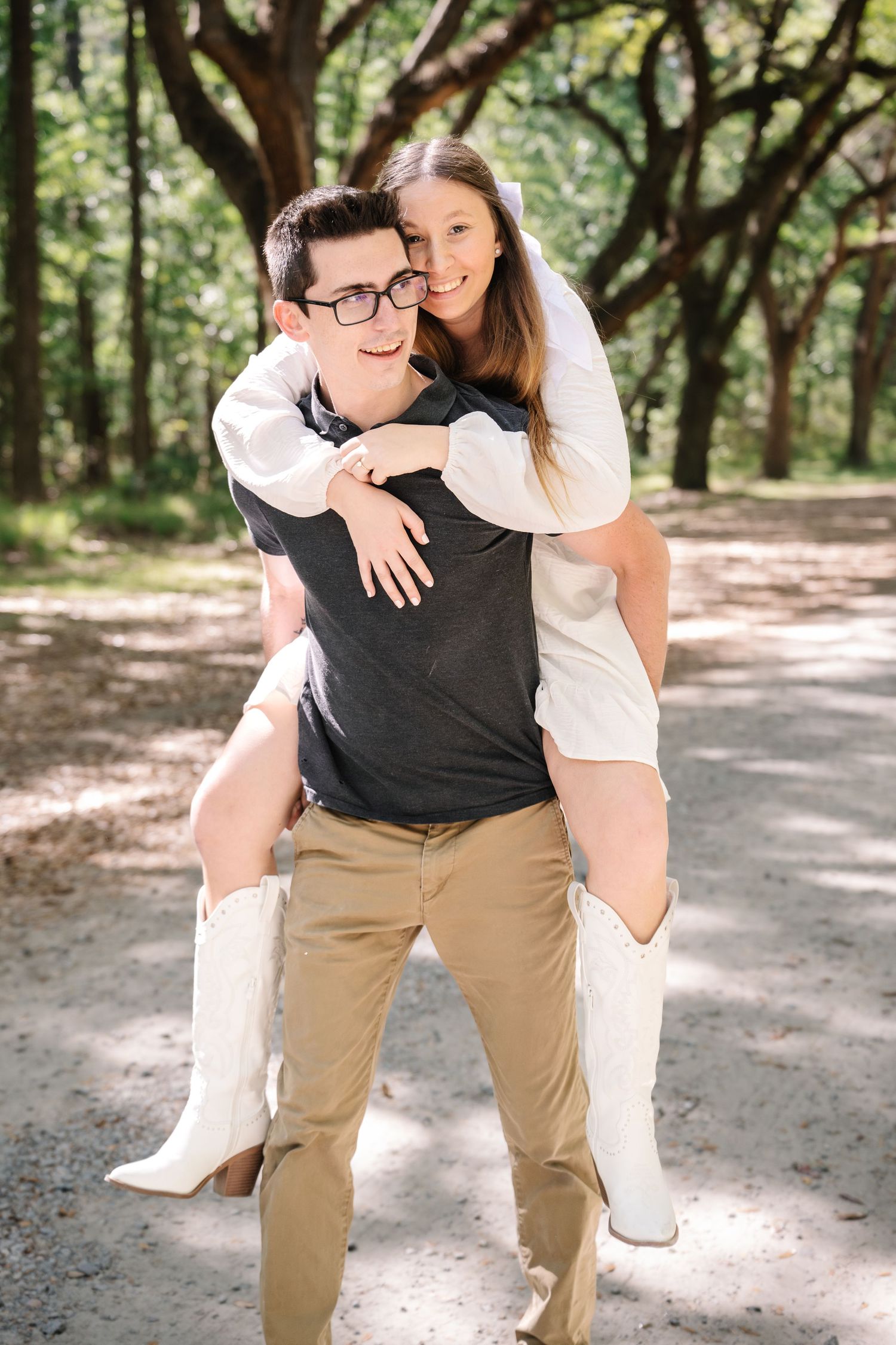 Man giving woman a piggyback ride along a sunlit tree-lined path, both smiling happily.