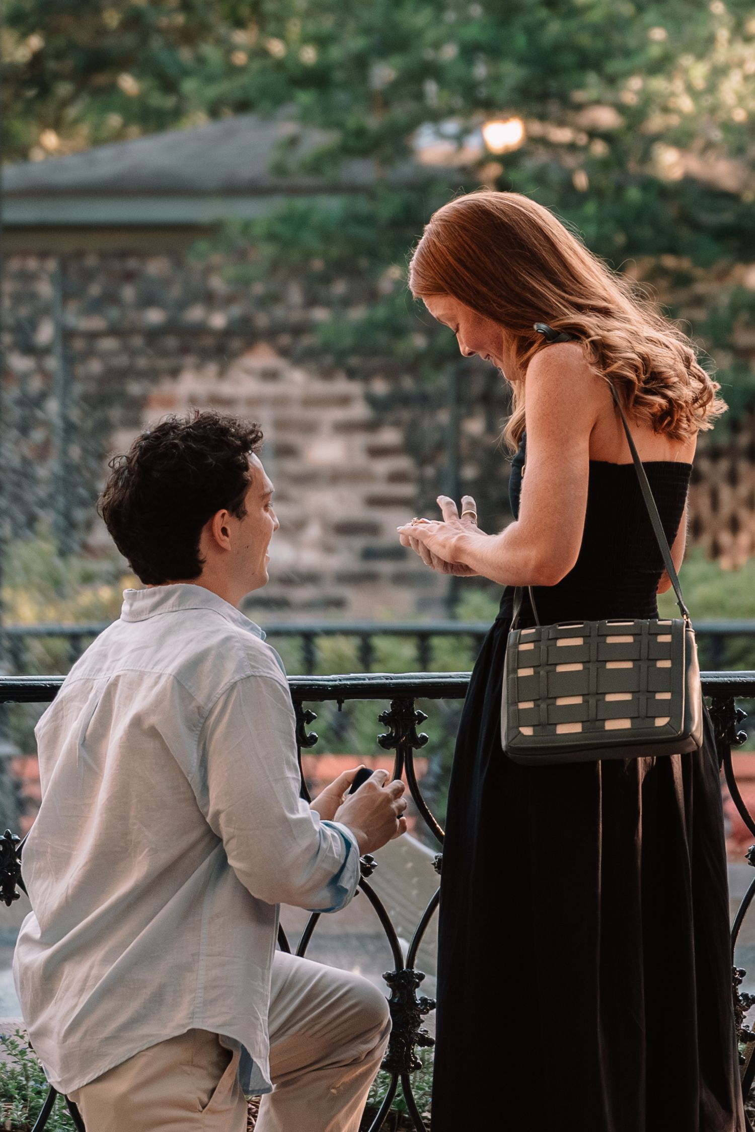 Man kneeling on one knee proposing to woman in black dress near ornate iron fence outdoors.