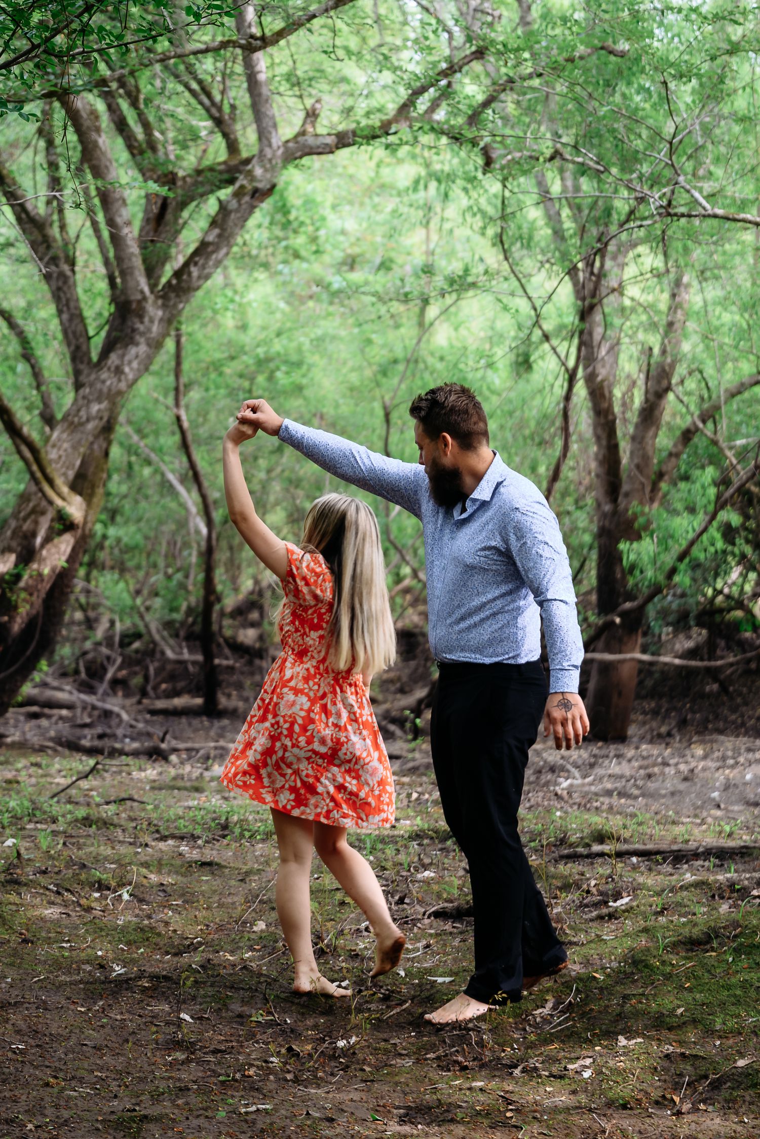 A bearded man in a blue shirt twirls a woman in a red floral dress barefoot in a lush green forest.
