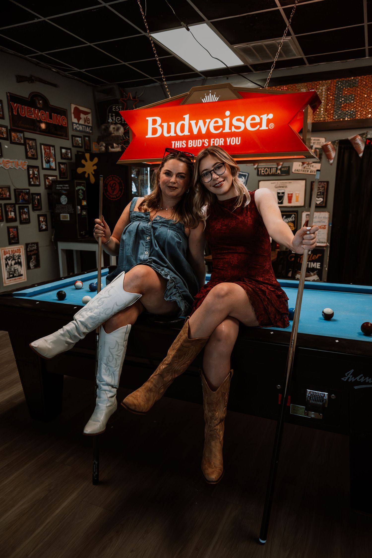 Two women posing under a Budweiser sign near a pool table in a bar.