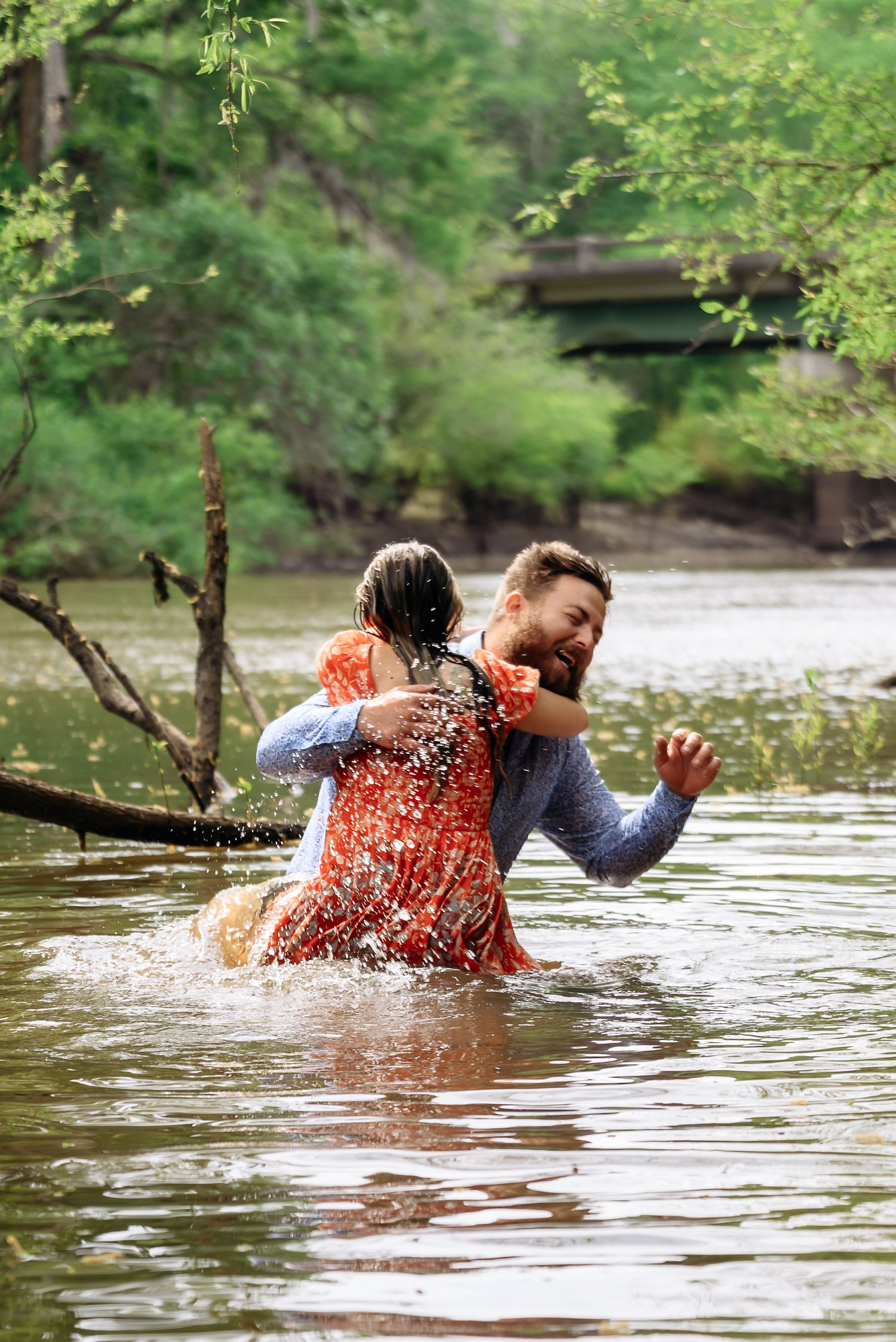 Couple laughing and splashing in a shallow river surrounded by lush green trees.