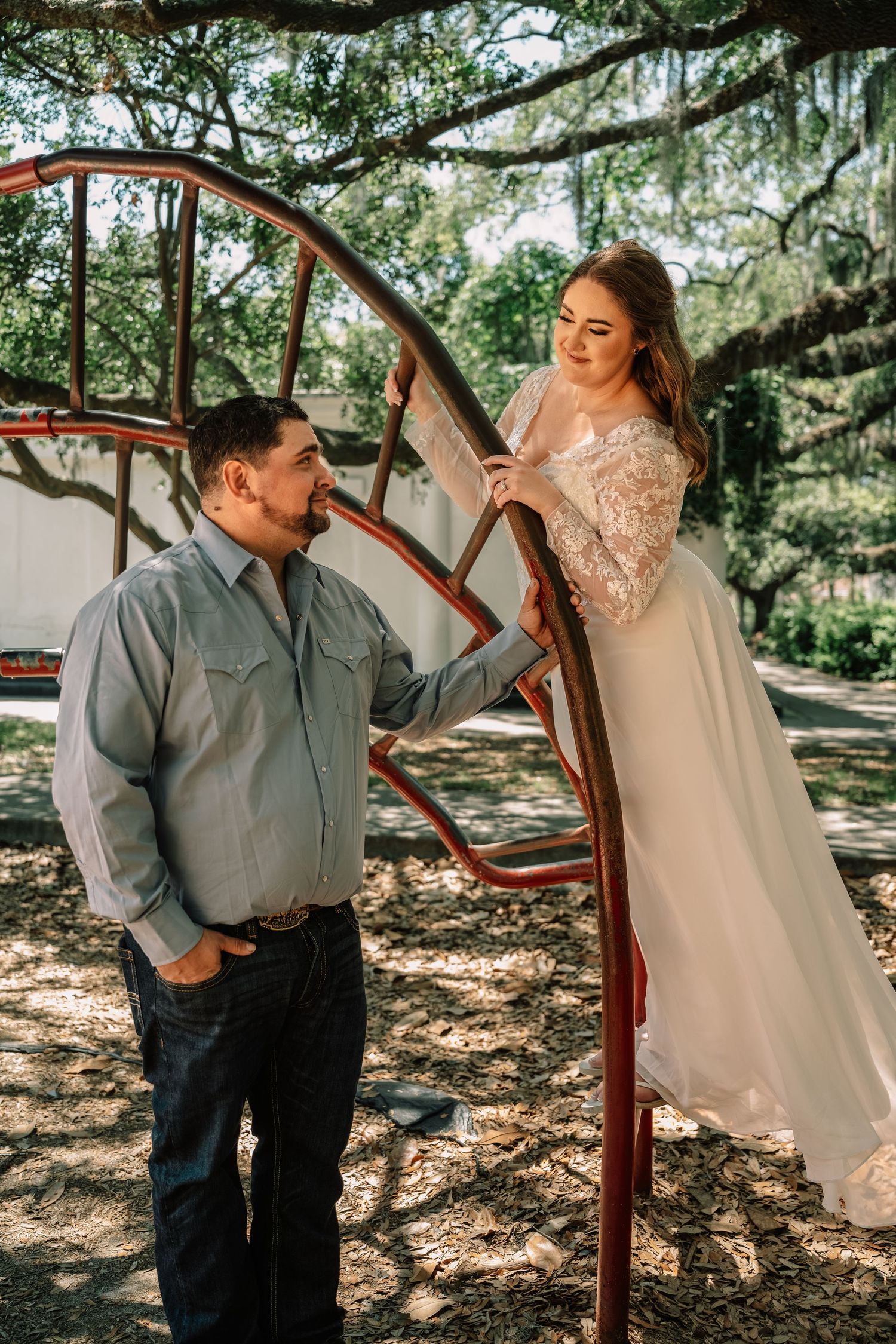Bride in lace gown smiles down at groom from a rustic red climbing structure beneath sprawling oak trees.