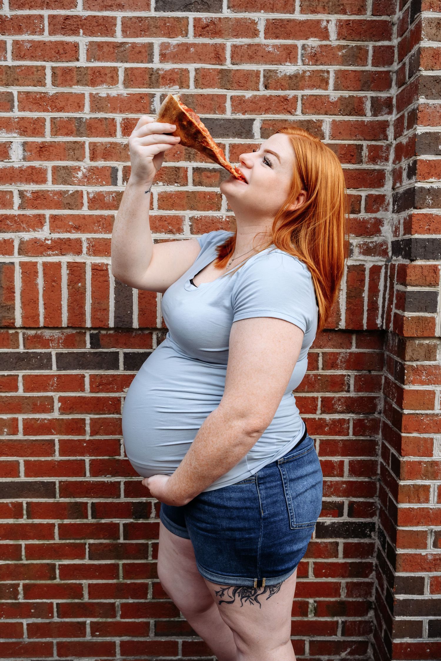 Pregnant redhead woman eating a slice of pizza while standing against a red brick wall outdoors.