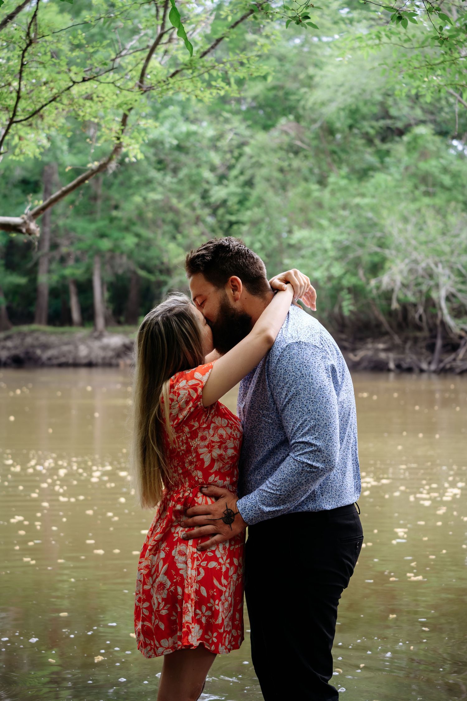 Couple sharing a romantic kiss by a serene river surrounded by lush green trees, woman in red floral dress.
