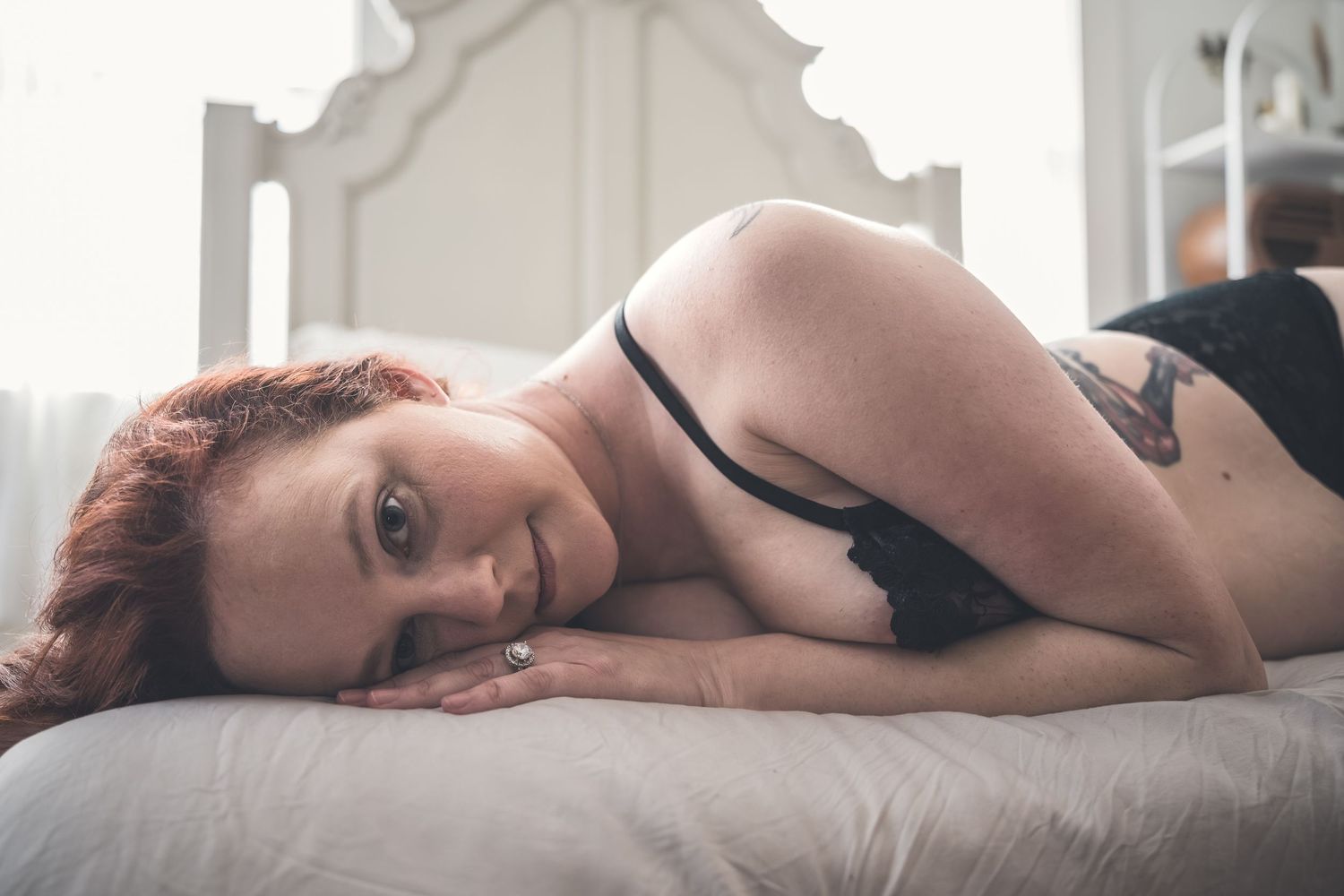 Redhead woman in black lingerie lying on white bed, looking at camera in intimate boudoir portrait photography.