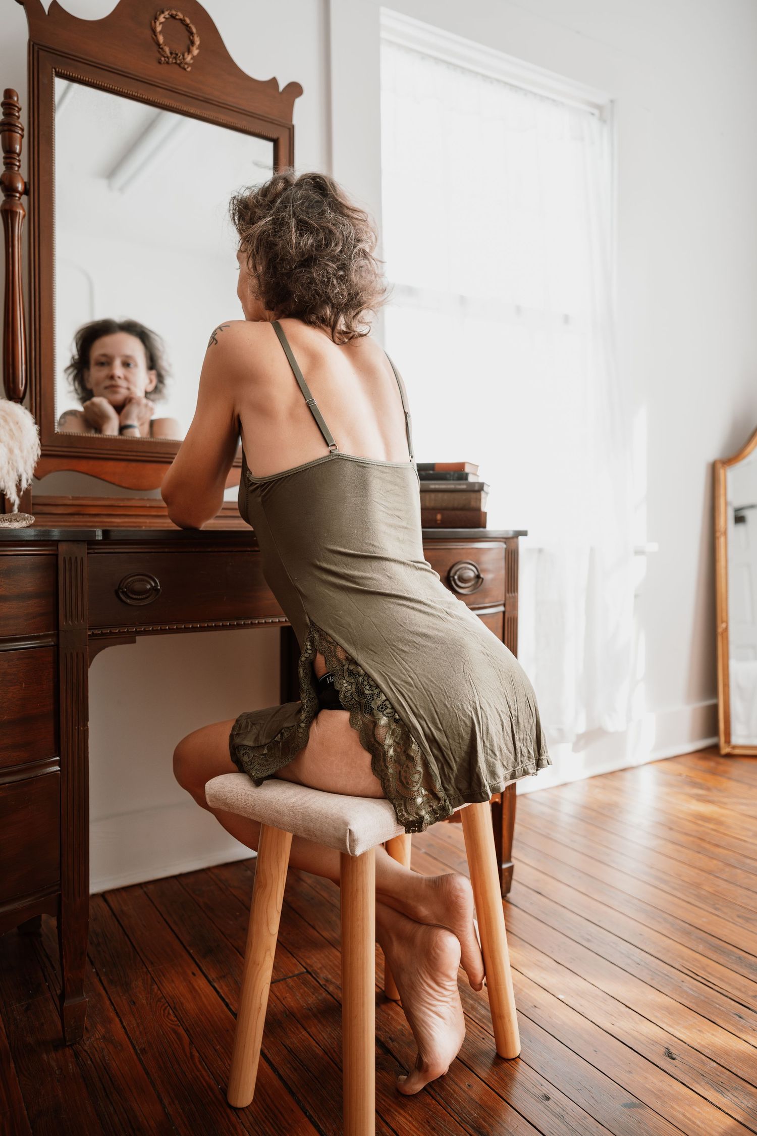 Woman in olive slip dress sits at antique vanity mirror on hardwood floor.