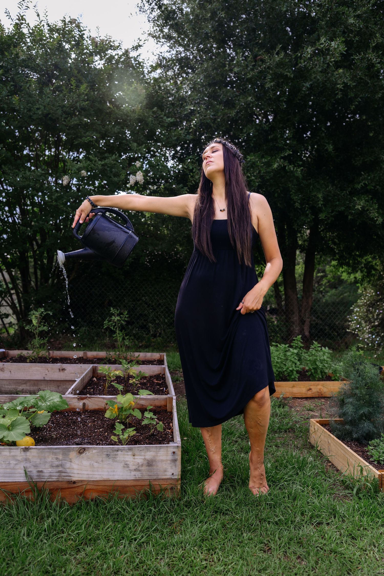 Barefoot woman in black dress watering raised garden beds with a black watering can.