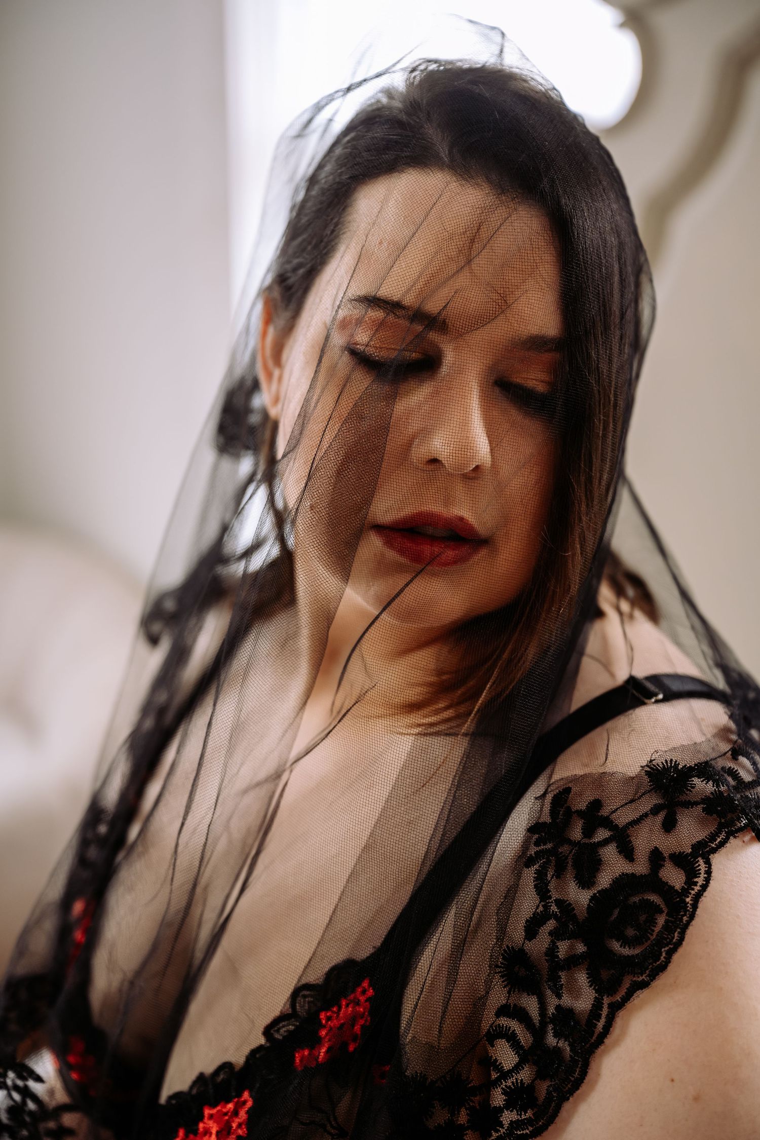 Woman wearing black lace lingerie and sheer black veil with eyes closed, soft natural window lighting portrait.