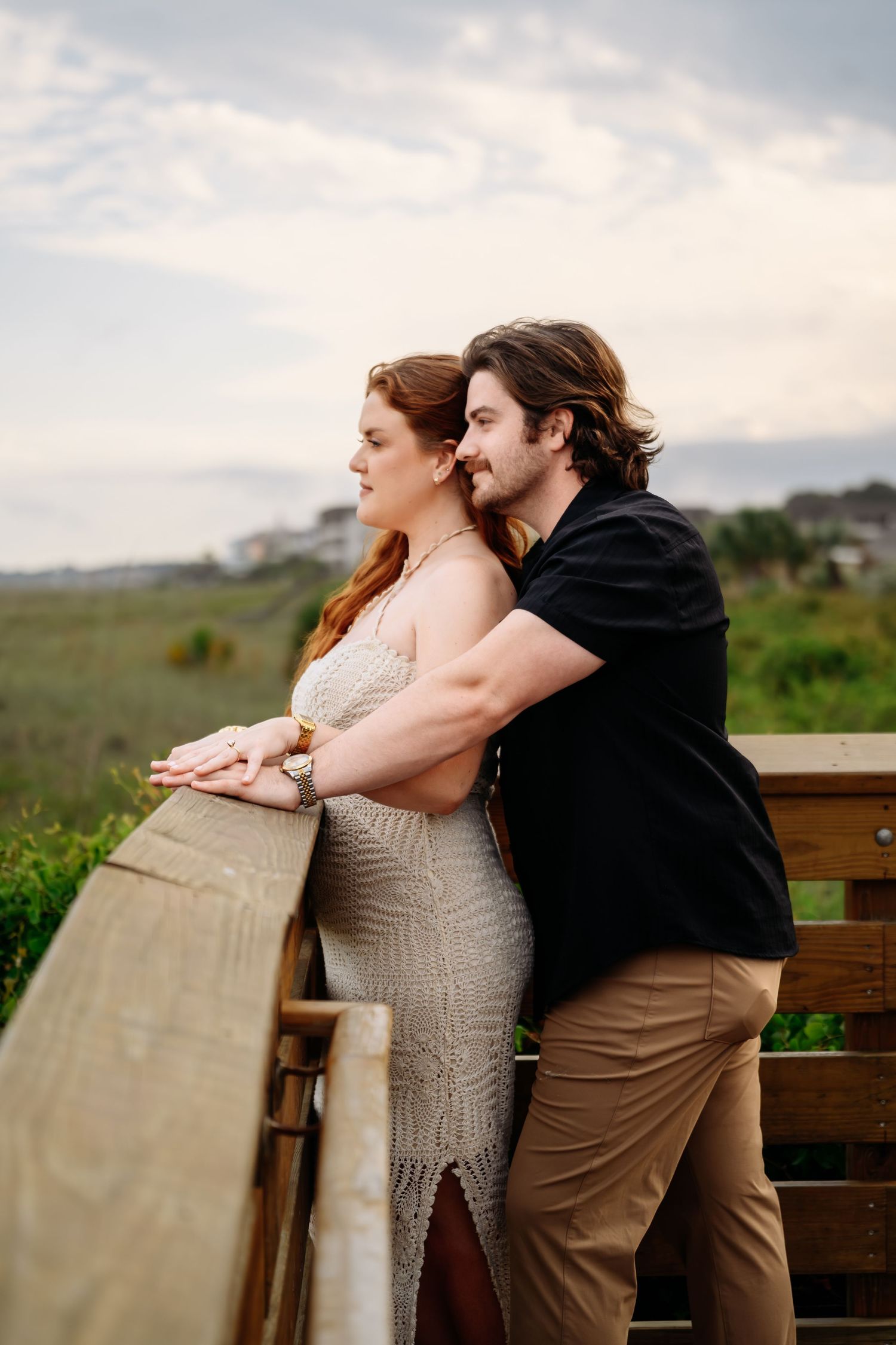 Engaged couple leaning on wooden railing overlooking coastal marsh at golden hour.