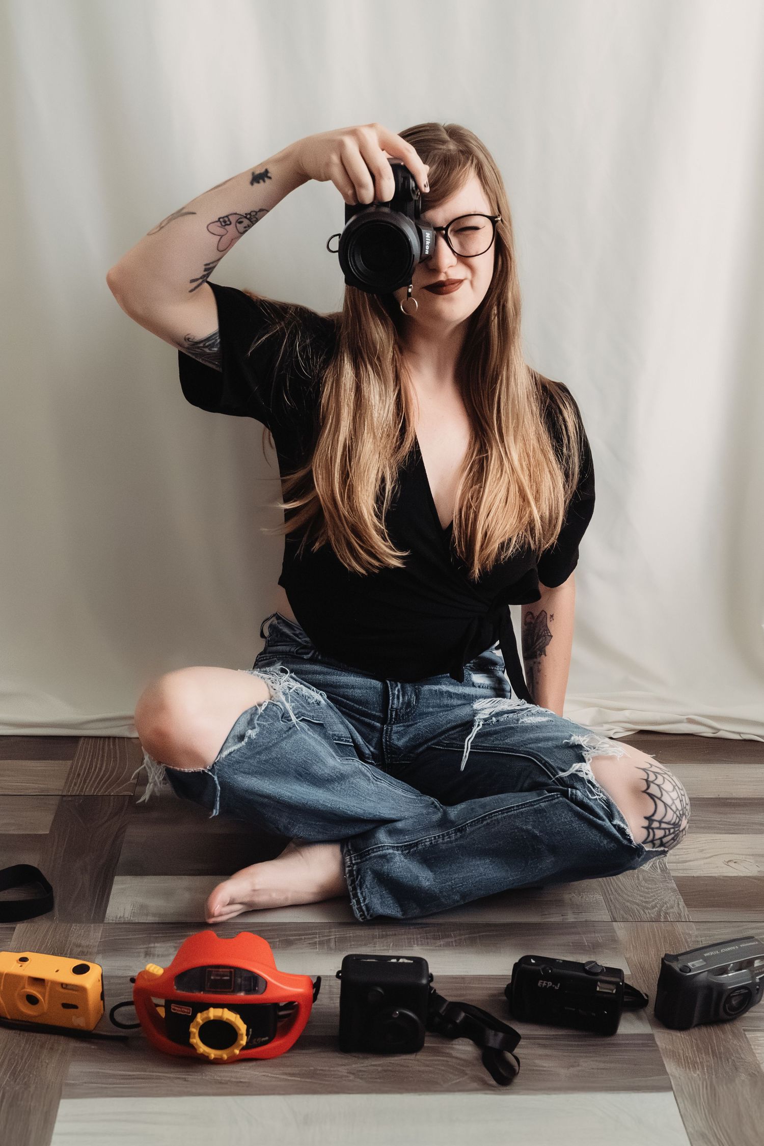 Tattooed female photographer sitting on floor pointing camera forward surrounded by vintage cameras.