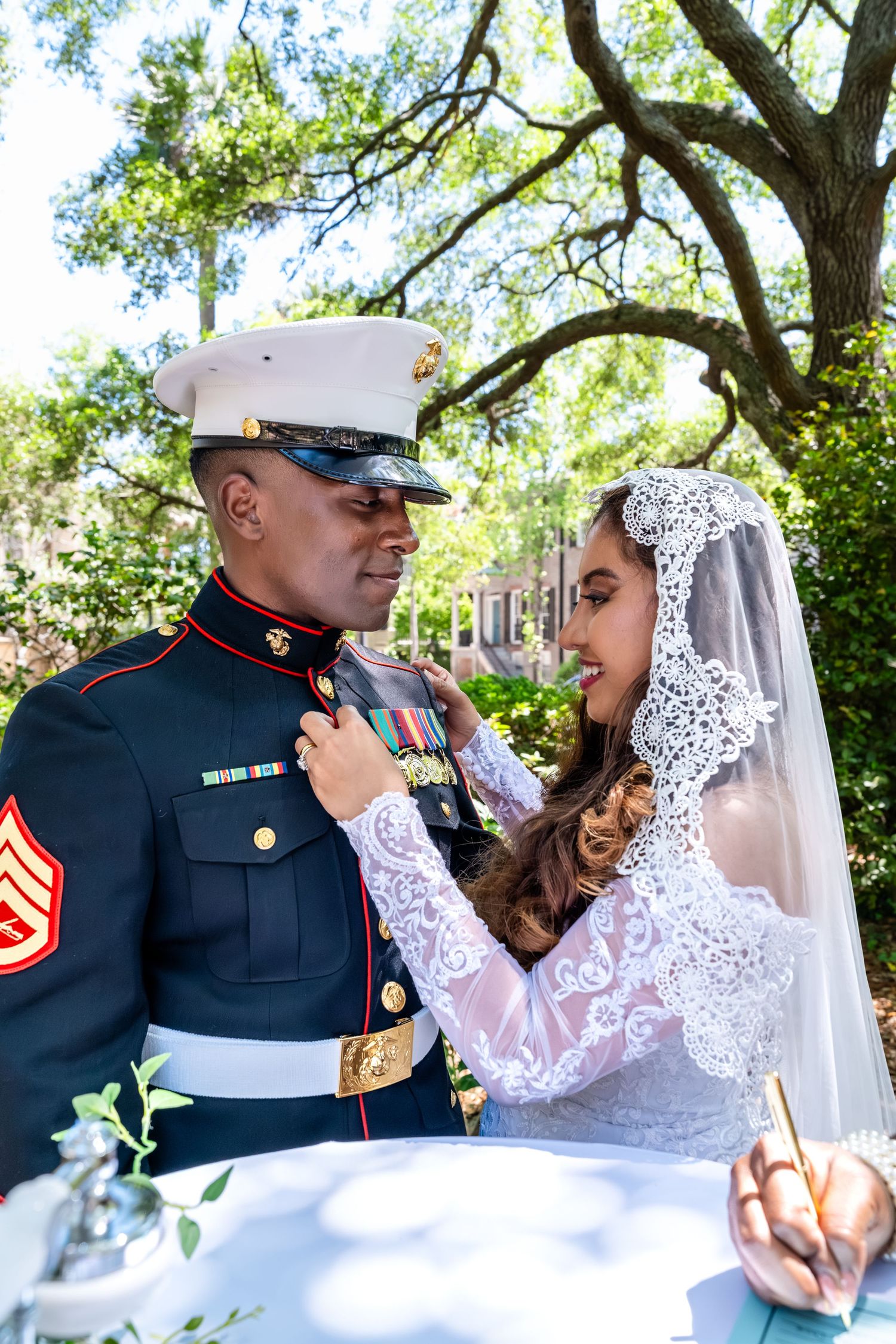Bride adjusts Marine groom's tie during outdoor wedding ceremony under shaded garden trees.