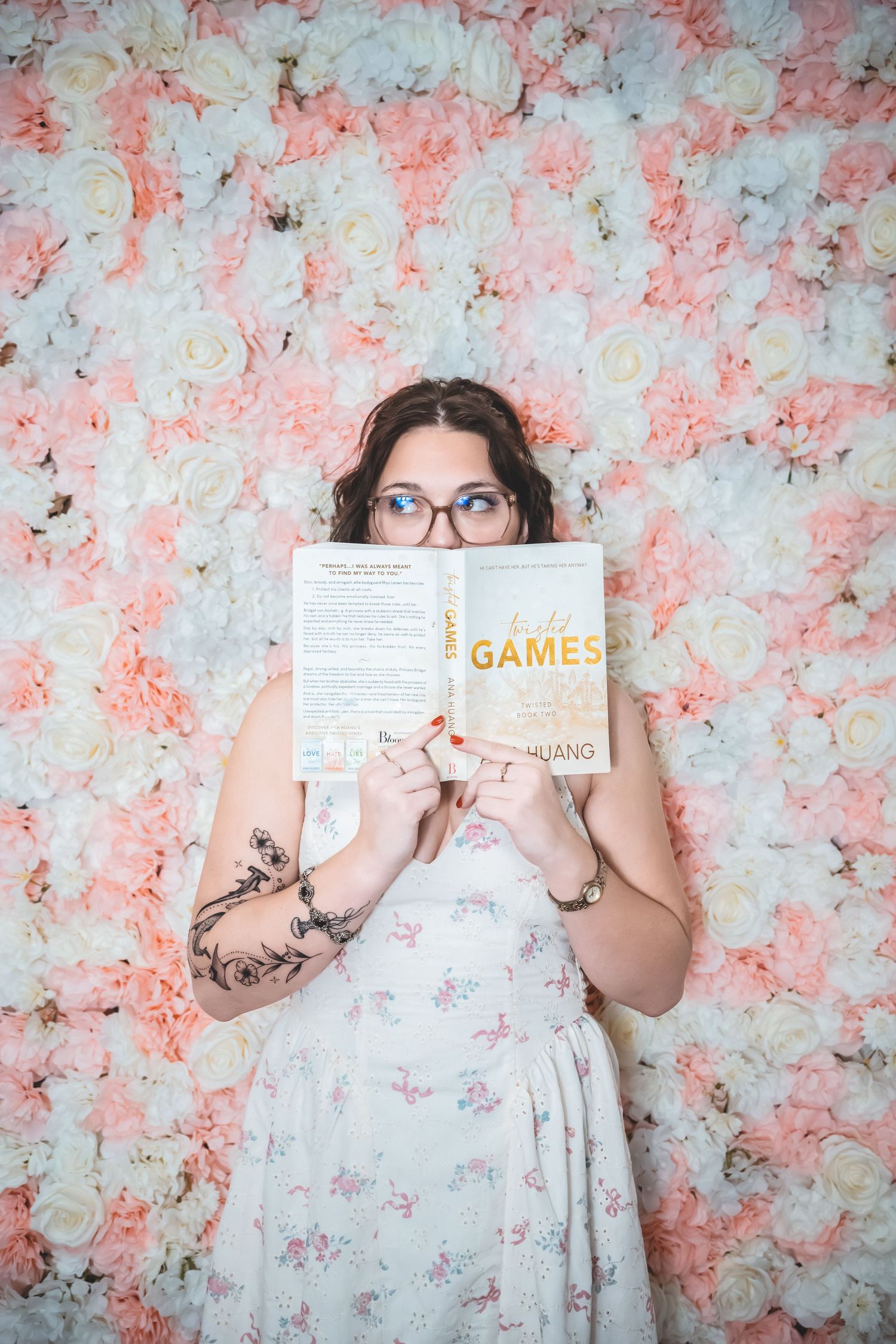 Someone in a floral dress poses with a book against a pink and white flower wall.