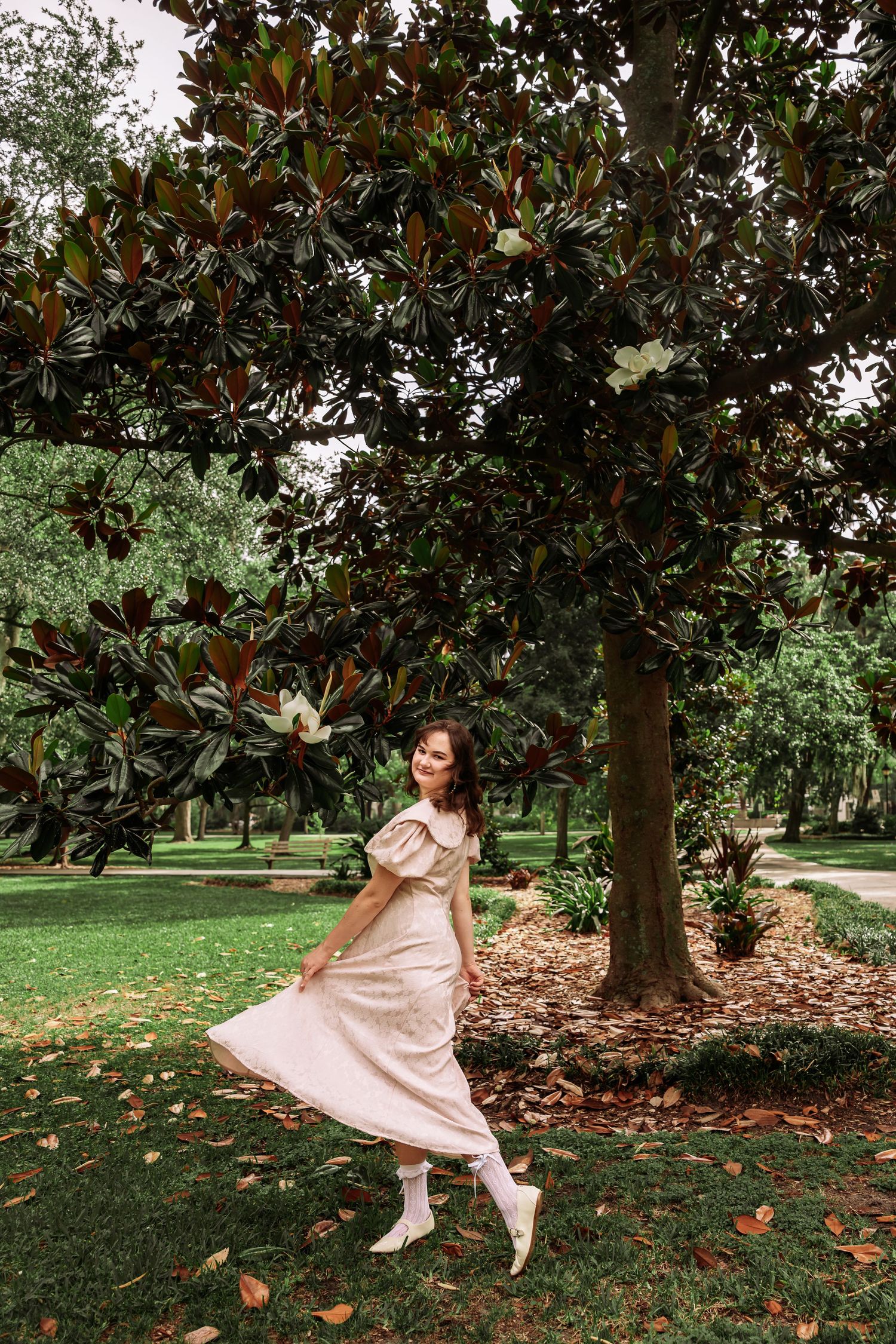 Woman in pink midi dress twirling beside a large magnolia tree in a lush green park.