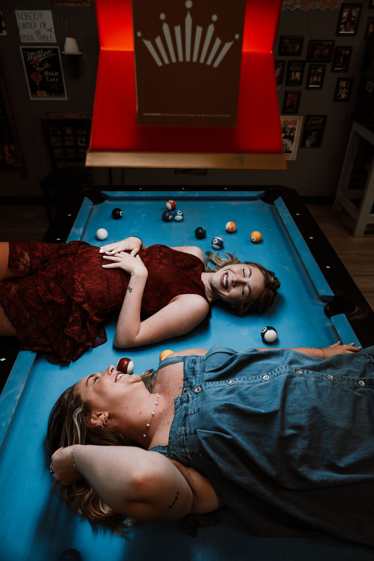 Two women laughing and lying on a blue pool table surrounded by colorful billiard balls indoors.