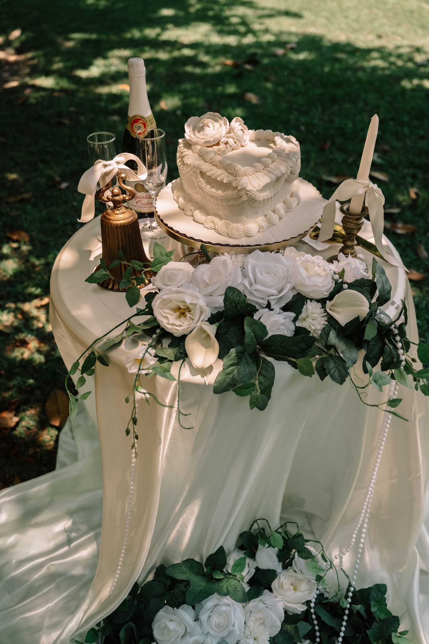 Elegant white wedding cake on a draped table surrounded by white roses and greenery in a garden setting.