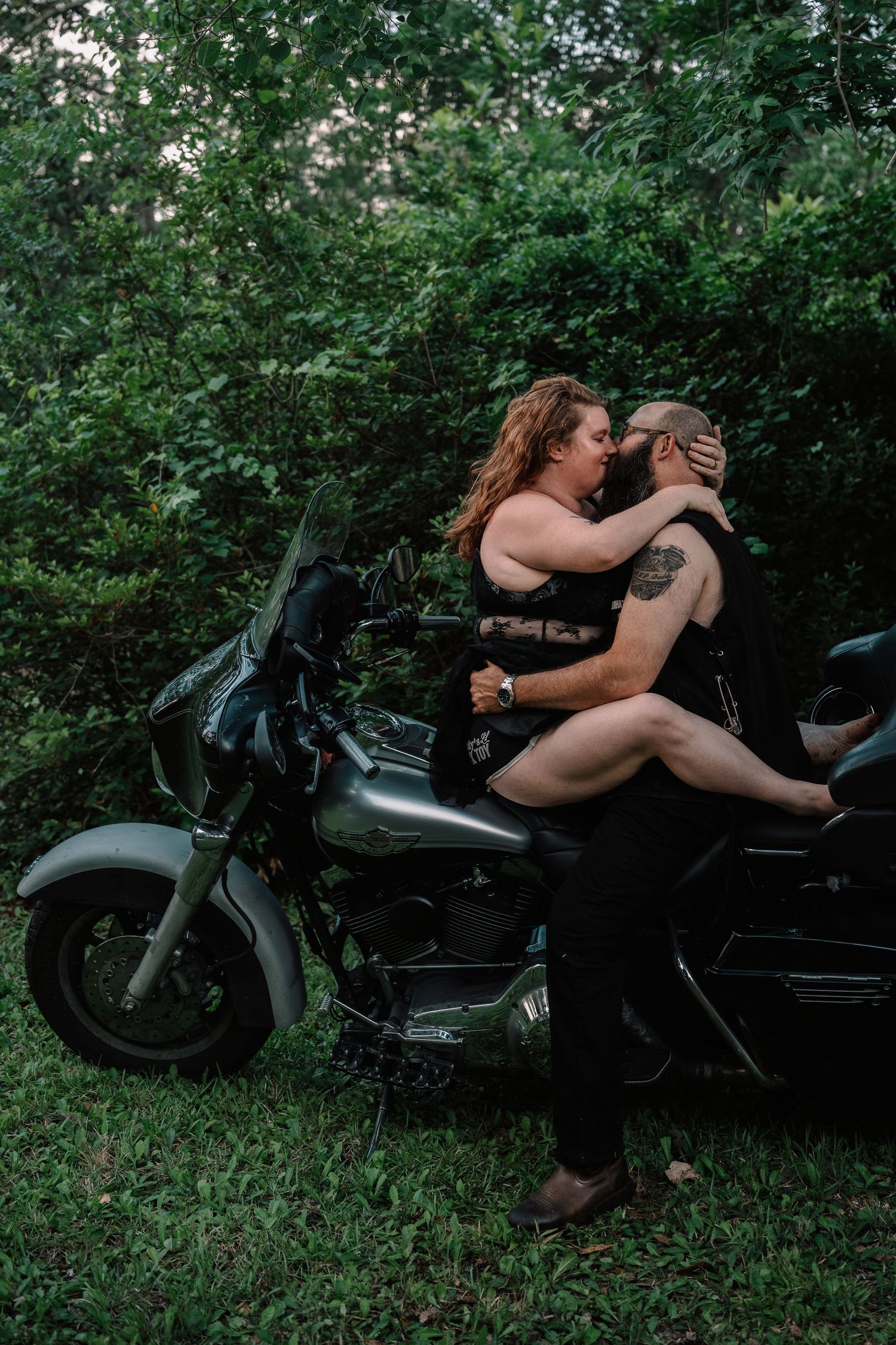 Couple sharing a romantic kiss beside a black motorcycle in a lush green forest setting.