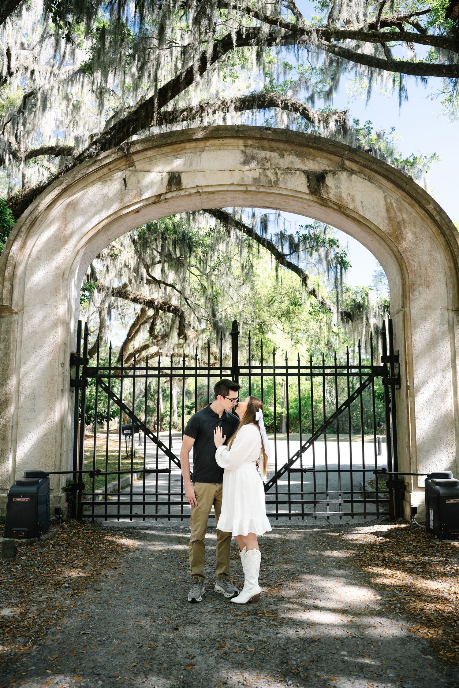 Couple sharing a kiss under a stone archway gate surrounded by moss-draped oak trees.