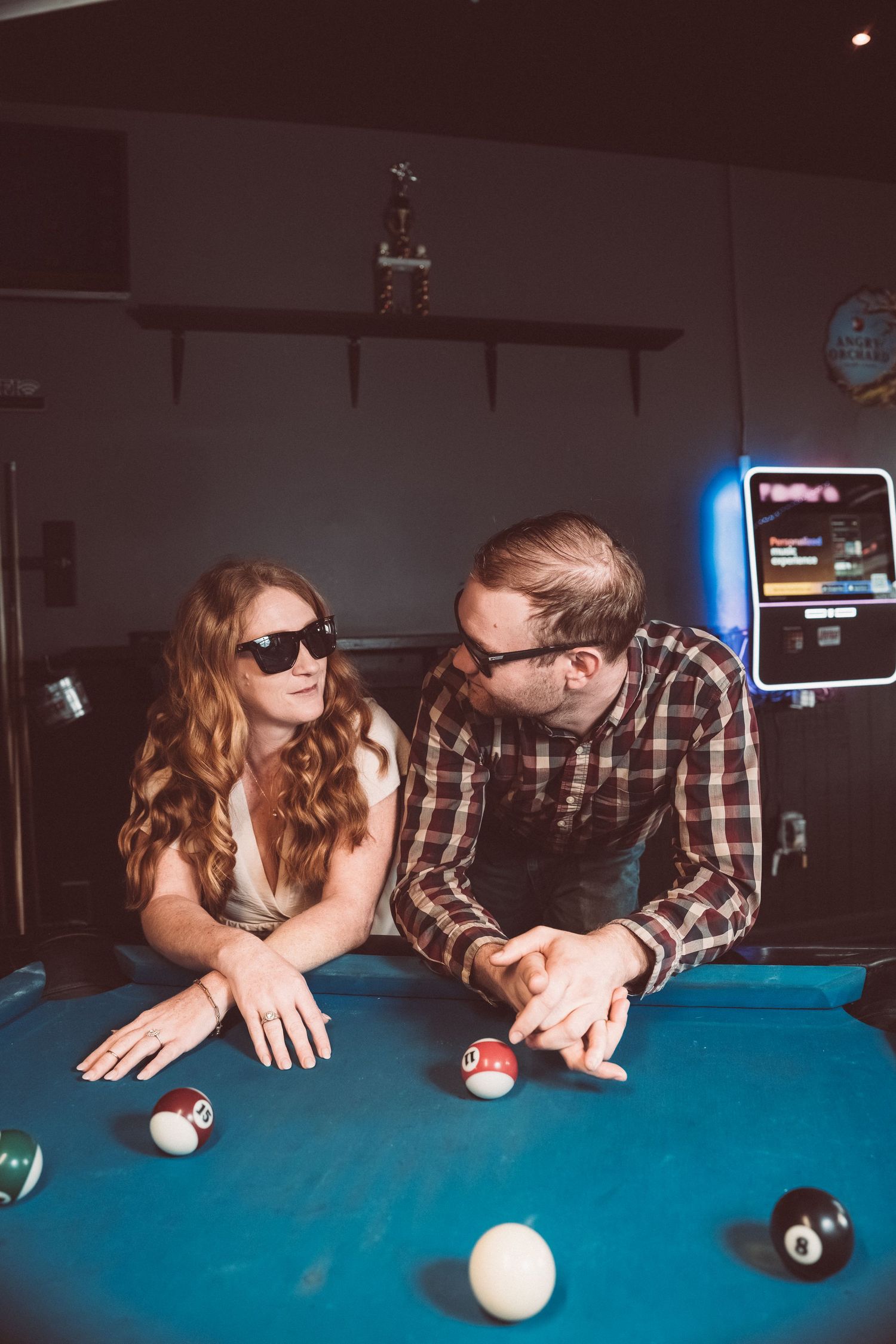 A playful couple leaning over a pool table wearing casual attire and sunglasses in a bar setting.