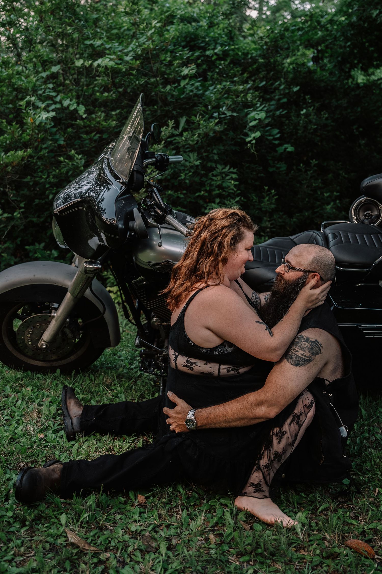 Couple sitting on grass sharing a tender moment beside a black motorcycle in nature.