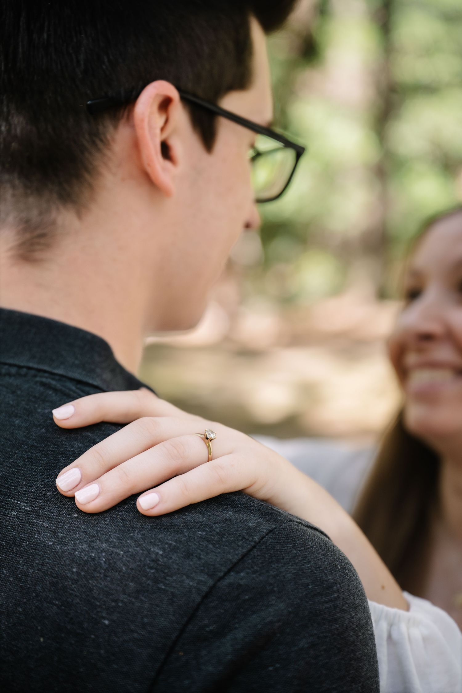 Close-up of womans hand with engagement ring resting on mans shoulder during outdoor photo session.
