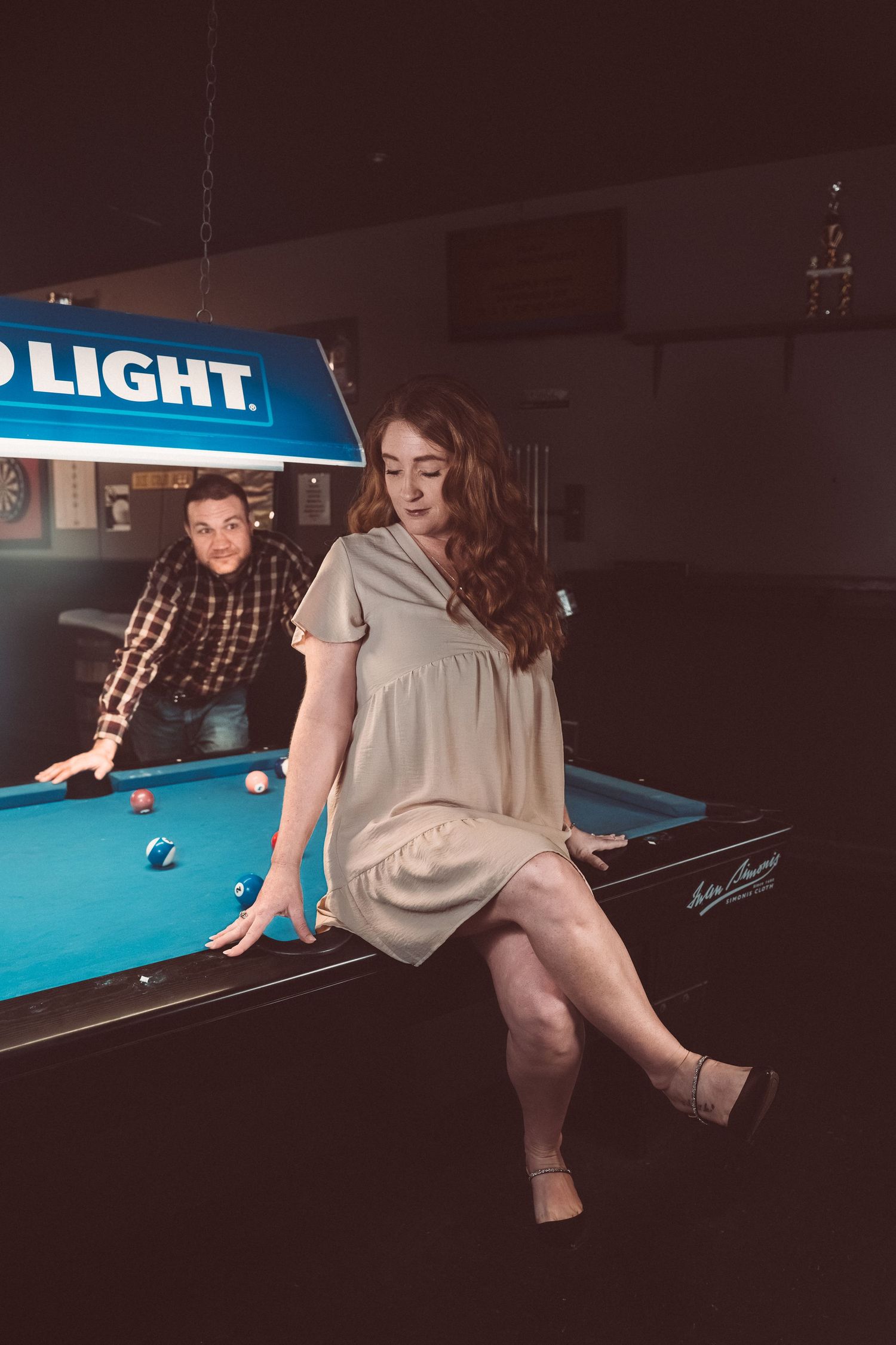 A couple plays pool at a dimly lit bar with a Bud Light sign overhead.