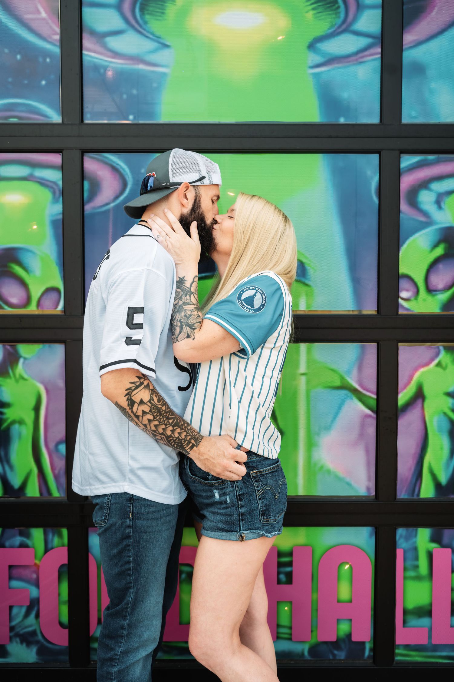 Tattooed couple sharing a kiss in front of colorful alien-themed graffiti wall wearing matching baseball jerseys.
