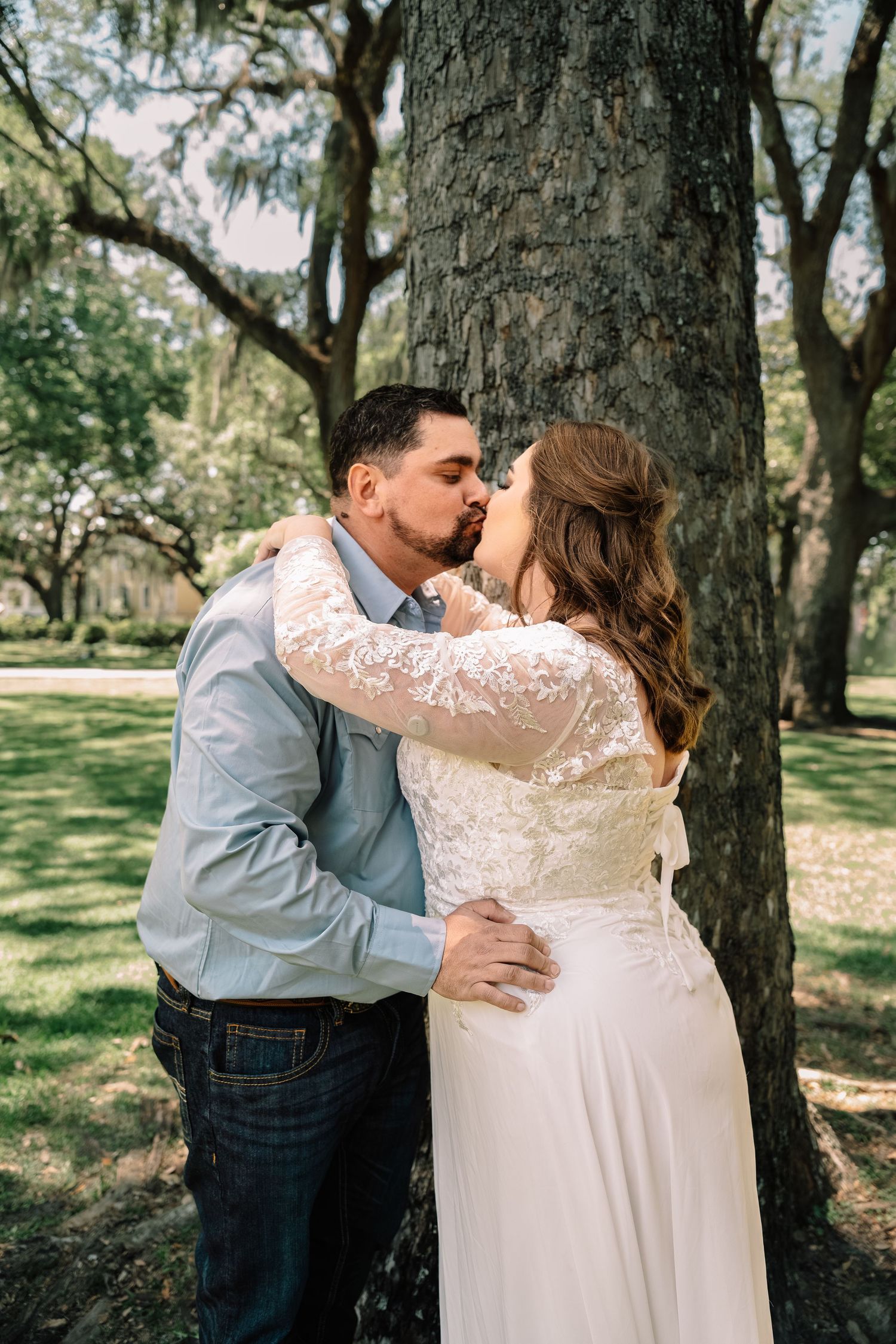 Newlywed couple shares a romantic kiss leaning against a large tree trunk in a sunlit park setting.