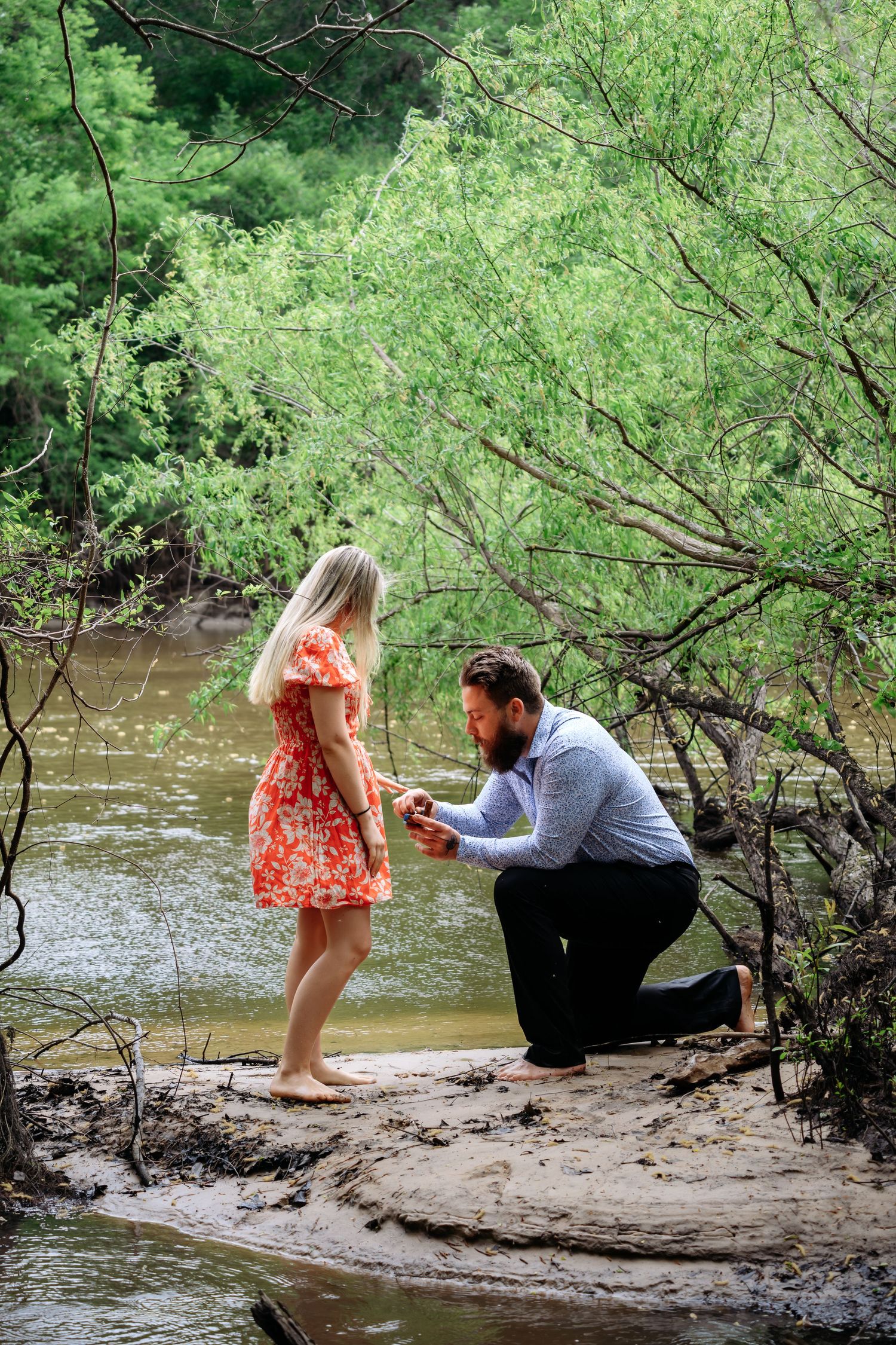 Bearded man kneels on one knee proposing to barefoot blonde woman in red dress beside a forest creek.