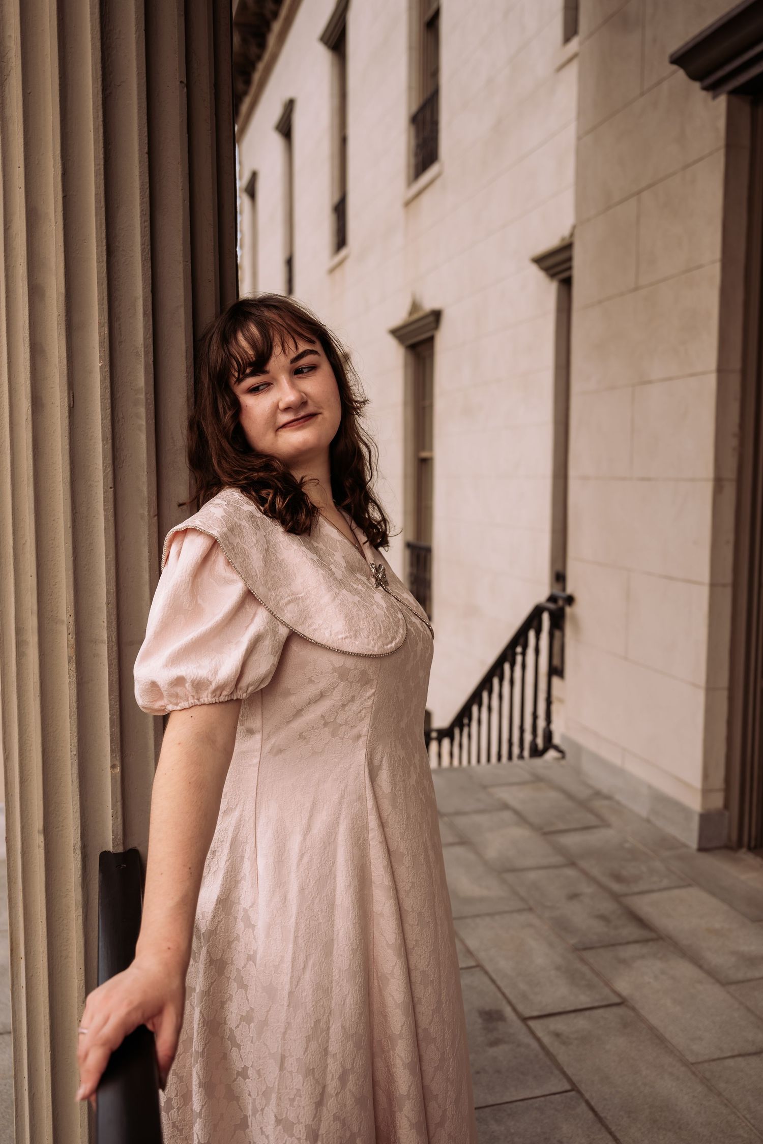 Young woman in blush pink puff sleeve dress leaning against stone columns of a building.