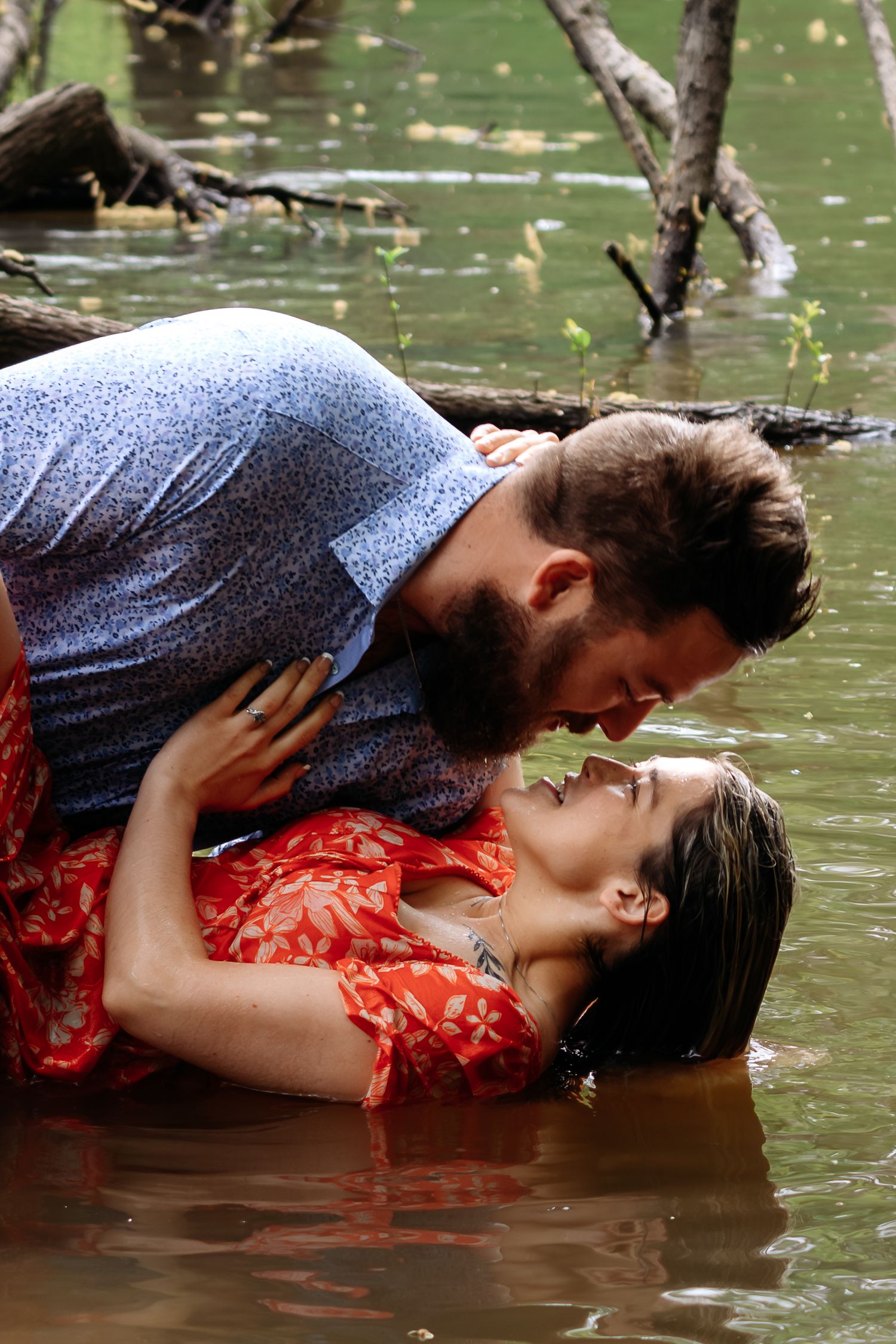 A bearded man in a blue shirt leans over a woman in a red floral dress lying in shallow water.