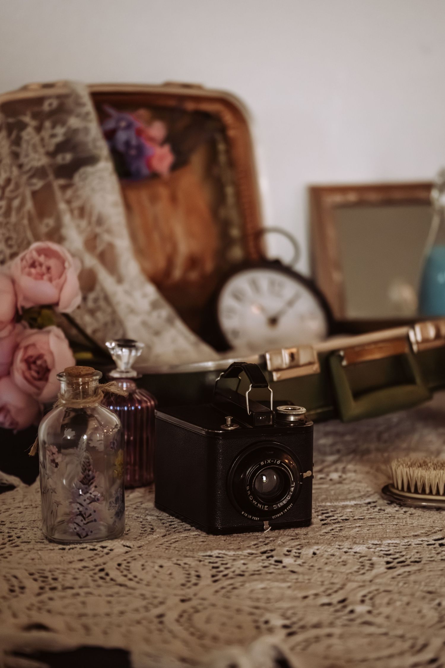 Vintage wedding flat lay with antique camera, perfume bottles, pink roses, clock, and lace on a table.
