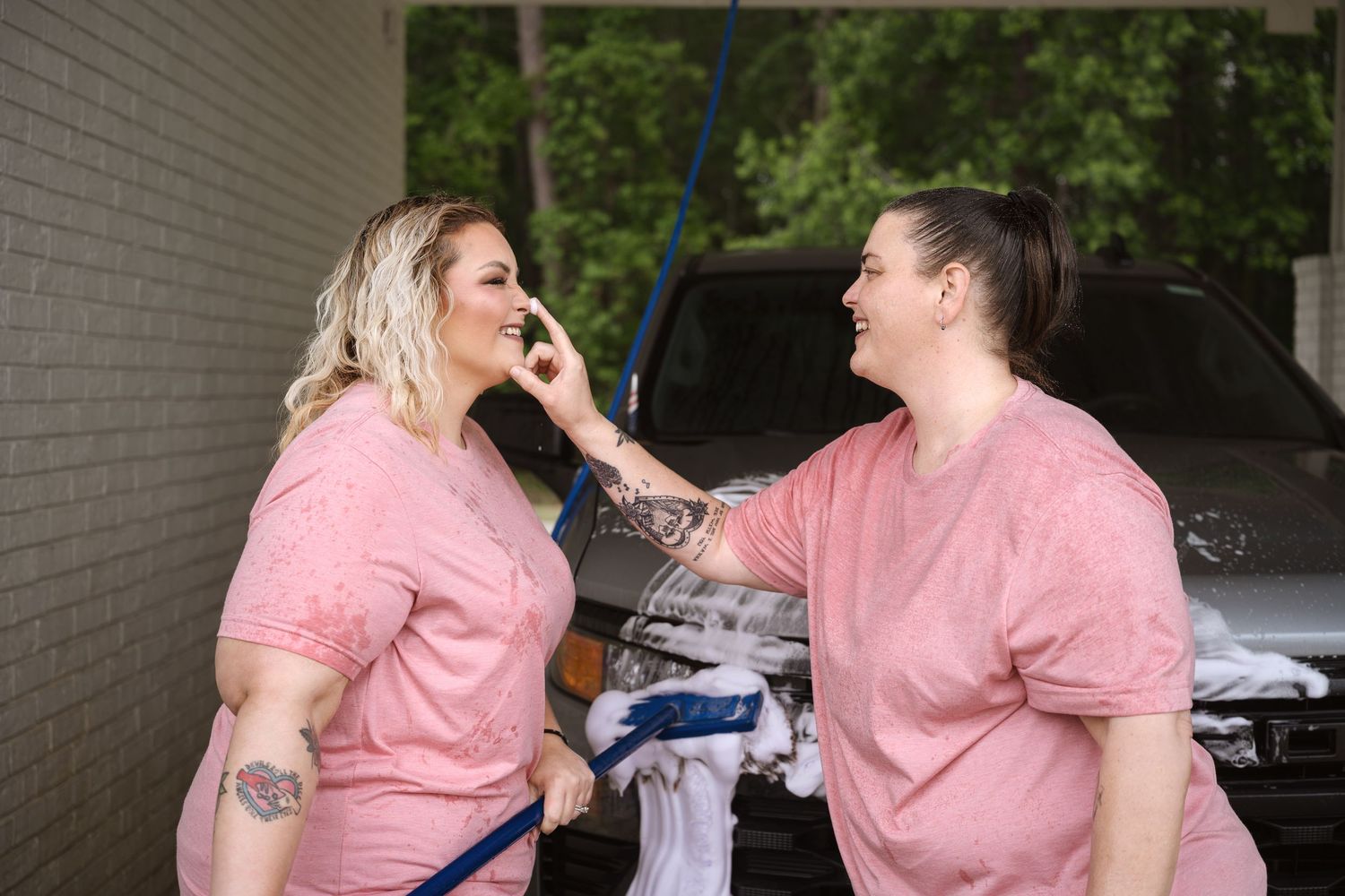 Two women in pink shirts laughing and playing with soap suds at a car wash.