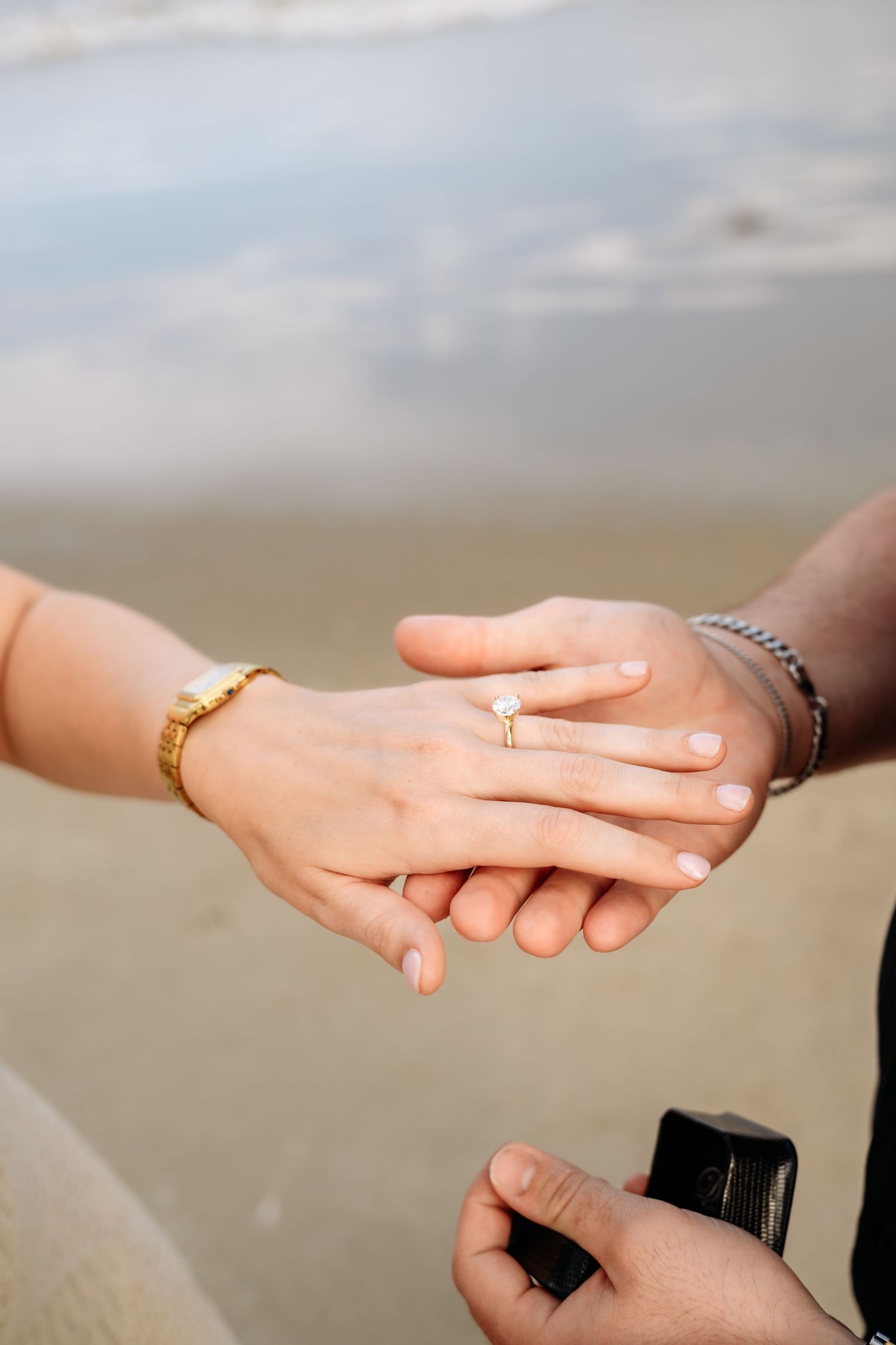 Close-up of hands showing engagement ring being placed on woman's finger at the beach.