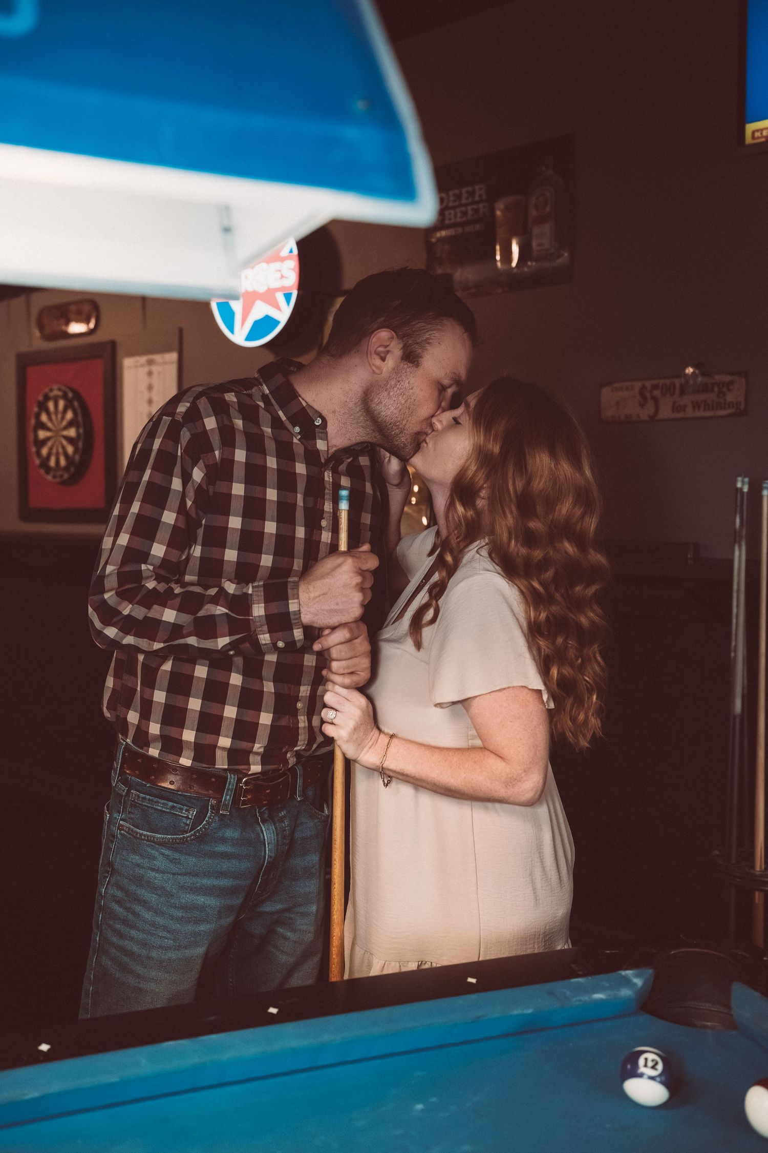 A couple shares a romantic moment while playing pool in a dimly lit bar with blue lighting overhead.