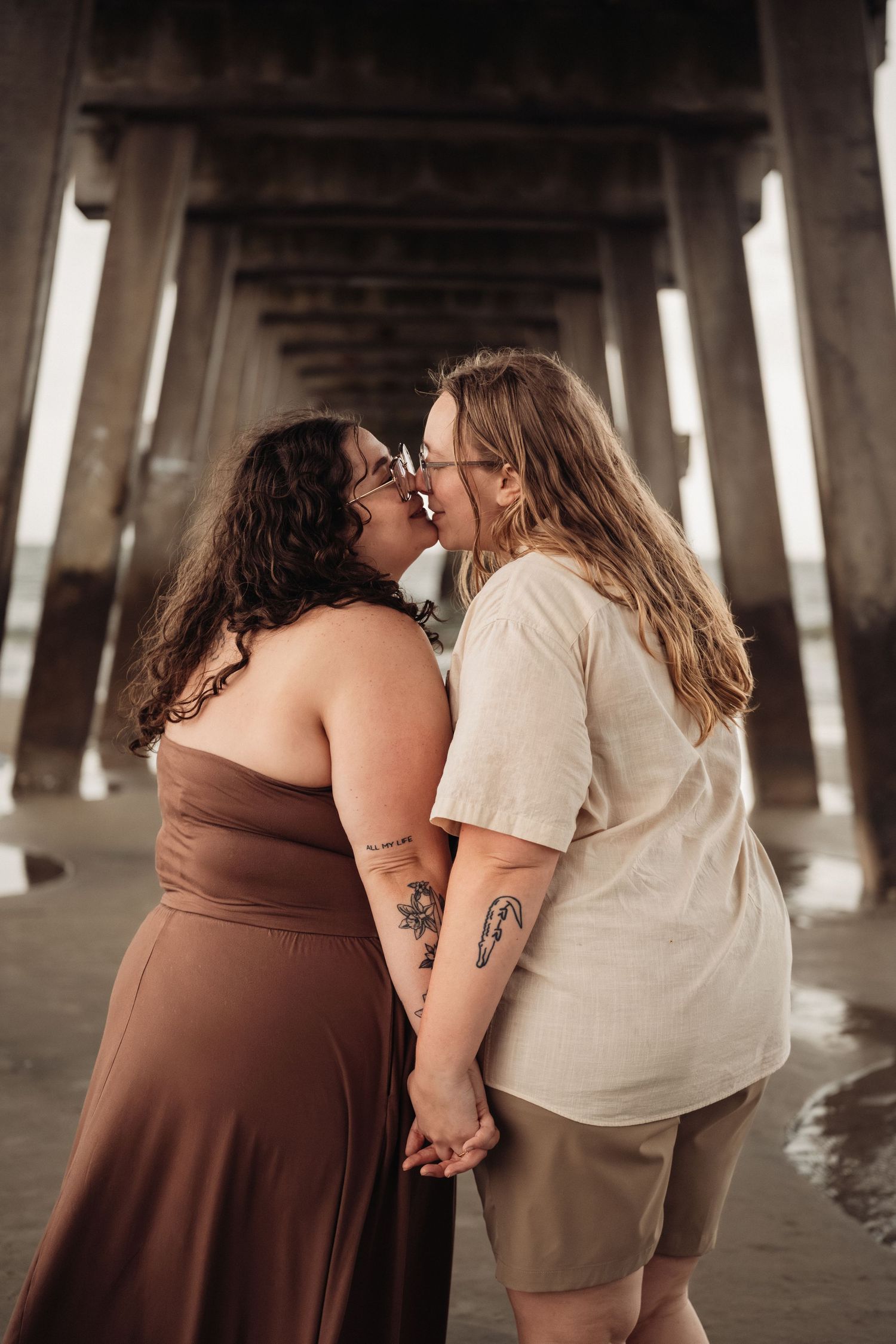 Two women sharing a romantic kiss while holding hands beneath a wooden beach pier structure.
